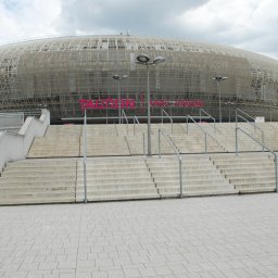 METAL-STYL Sp. z o.o. - Szerokie, betonowe schody z metalowymi poręczami prowadzące do nowoczesnego stadionu z siatkową konstrukcją elewacji i dużym, różowym napisem na fasadzie.