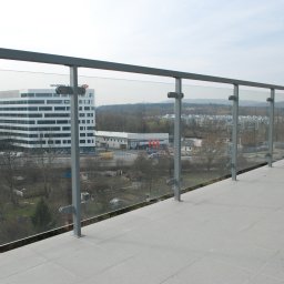 METAL-STYL Sp. z o.o. - Balkon z szarą, metalową balustradą i szklanym wypełnieniem, widok na budynki biurowe i okolicę.