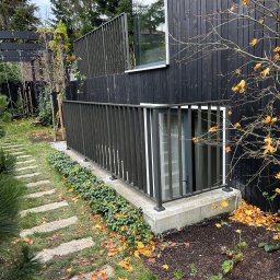 METAL-STYL Sp. z o.o. - Ciemna, minimalistyczna balustrada metalowa przy oknie piwnicznym, otoczona zielenią i opadłymi liśćmi, z betonową ścieżką w tle.