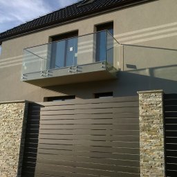 METAL-STYL Sp. z o.o. - Nowoczesny balkon z szklaną balustradą mocowaną na metalowych wspornikach, widok z dołu na tle domu z szarą elewacją i metalowym ogrodzeniem z elementami kamiennymi.