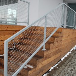 METAL-STYL Sp. z o.o. - Zewnętrzne drewniane schody z siatkową balustradą w kolorze jasnoszarym, prowadzące do budynku z szarą bramą garażową i drewnianą elewacją, na ziemi resztki śniegu.