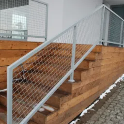 Zewnętrzne drewniane schody z siatkową balustradą w kolorze jasnoszarym, prowadzące do budynku z szarą bramą garażową i drewnianą elewacją, na ziemi resztki śniegu.