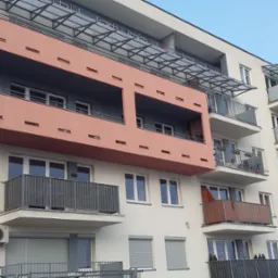 Nowoczesny budynek mieszkalny z balkonami zabezpieczonymi metalowymi barierkami, widoczne elementy konstrukcji zadaszenia nad balkonami na wyższych piętrach.