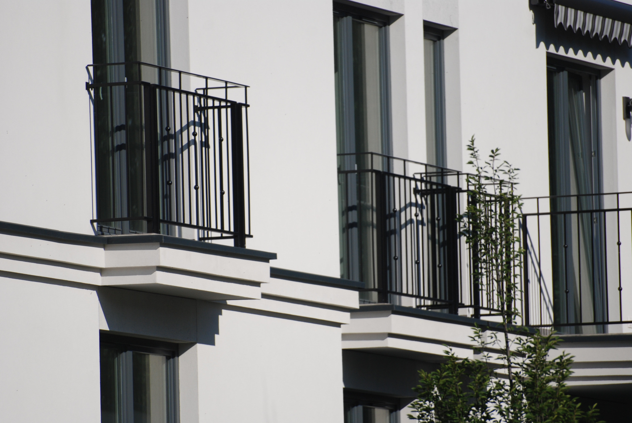 Elewacja budynku z trzema balkonami z czarnymi, metalowymi balustradami, zieleń drzewek widoczna na tle jednego z balkonów.
