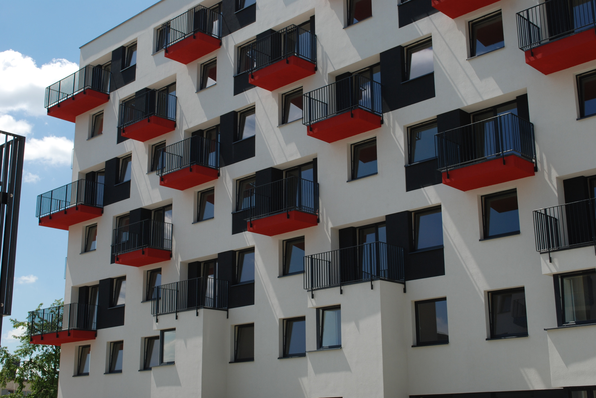 Fasada nowoczesnego budynku mieszkalnego z balkonami o czerwonych podłogach i czarnych, metalowych barierkach.
