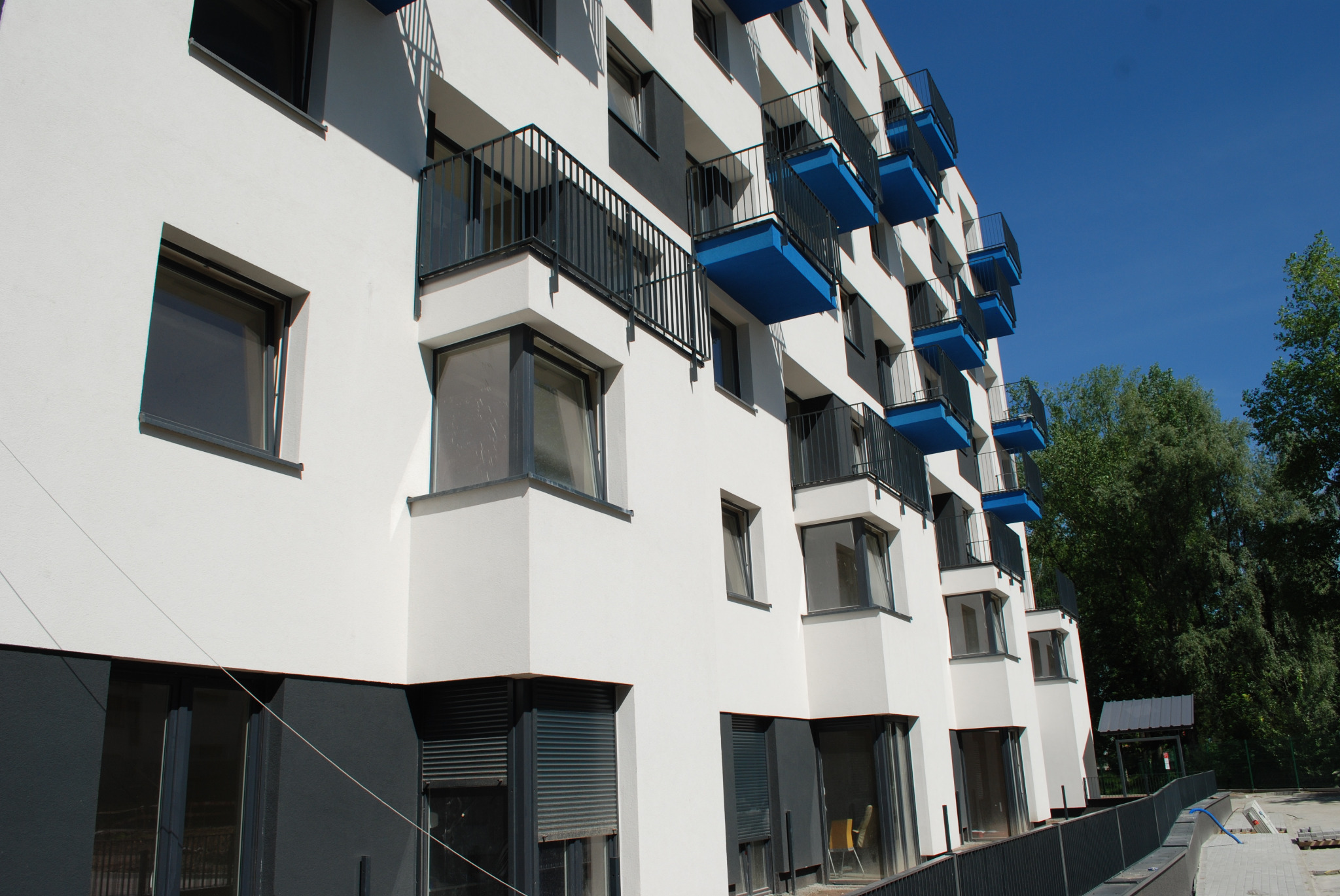 Fasada budynku z balkonami z czarnymi balustradami i niebieskim spodem, widok z dołu na tle błękitnego nieba i drzew
