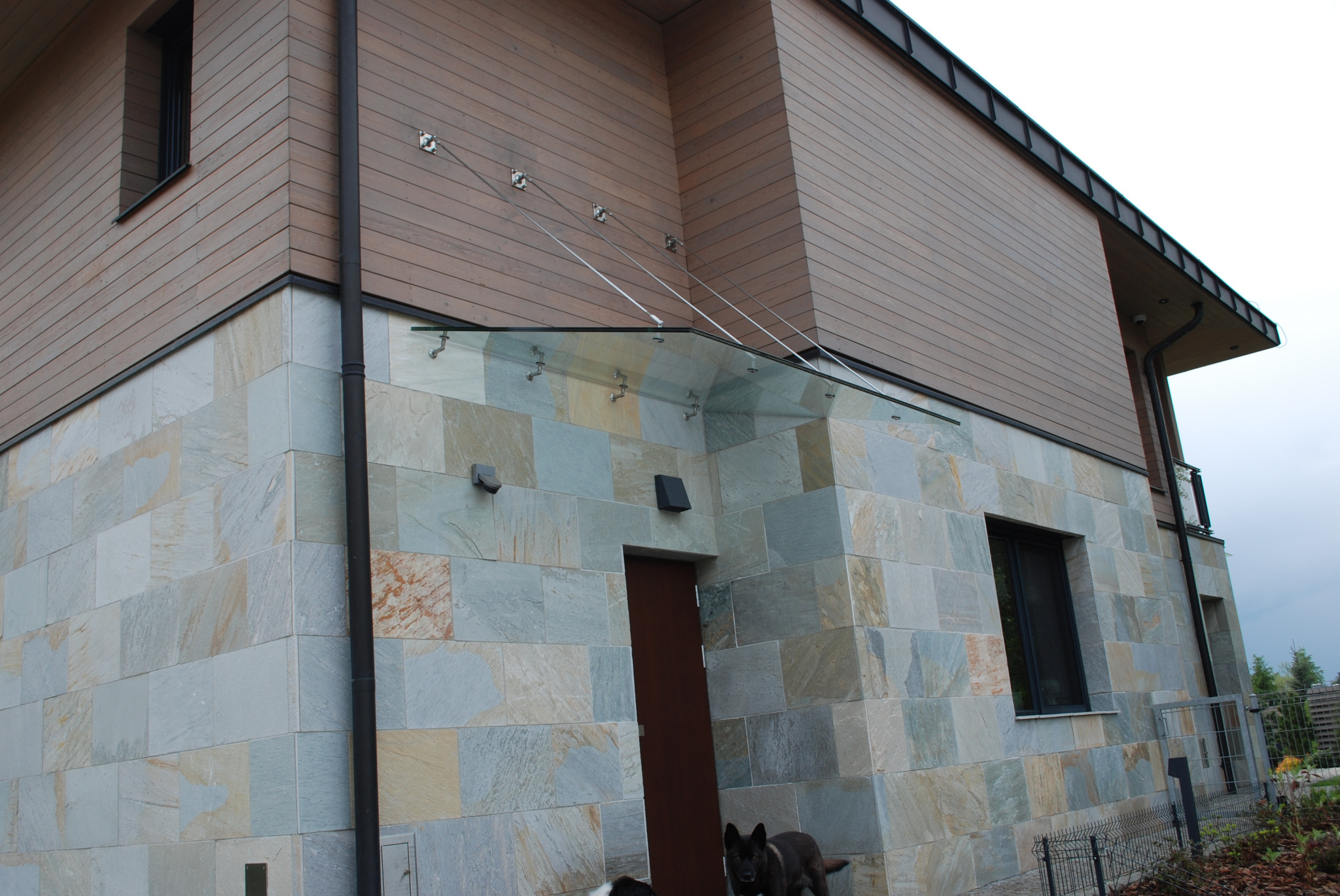 Szklany daszek nad wejściem do domu z elewacją z kamienia naturalnego, podtrzymywany przez stalowe linki, widoczne dwa psy.