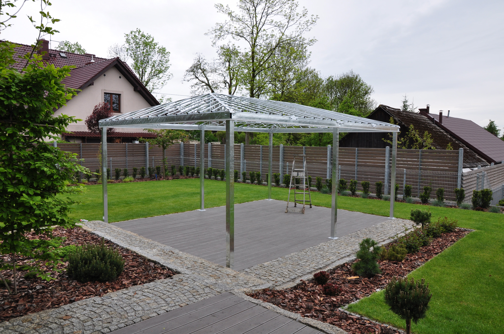 Konstrukcja metalowa altany z nierdzewnej stali na drewnianym podeście, widoczna drabina, w tle dom i ogrodzenie.