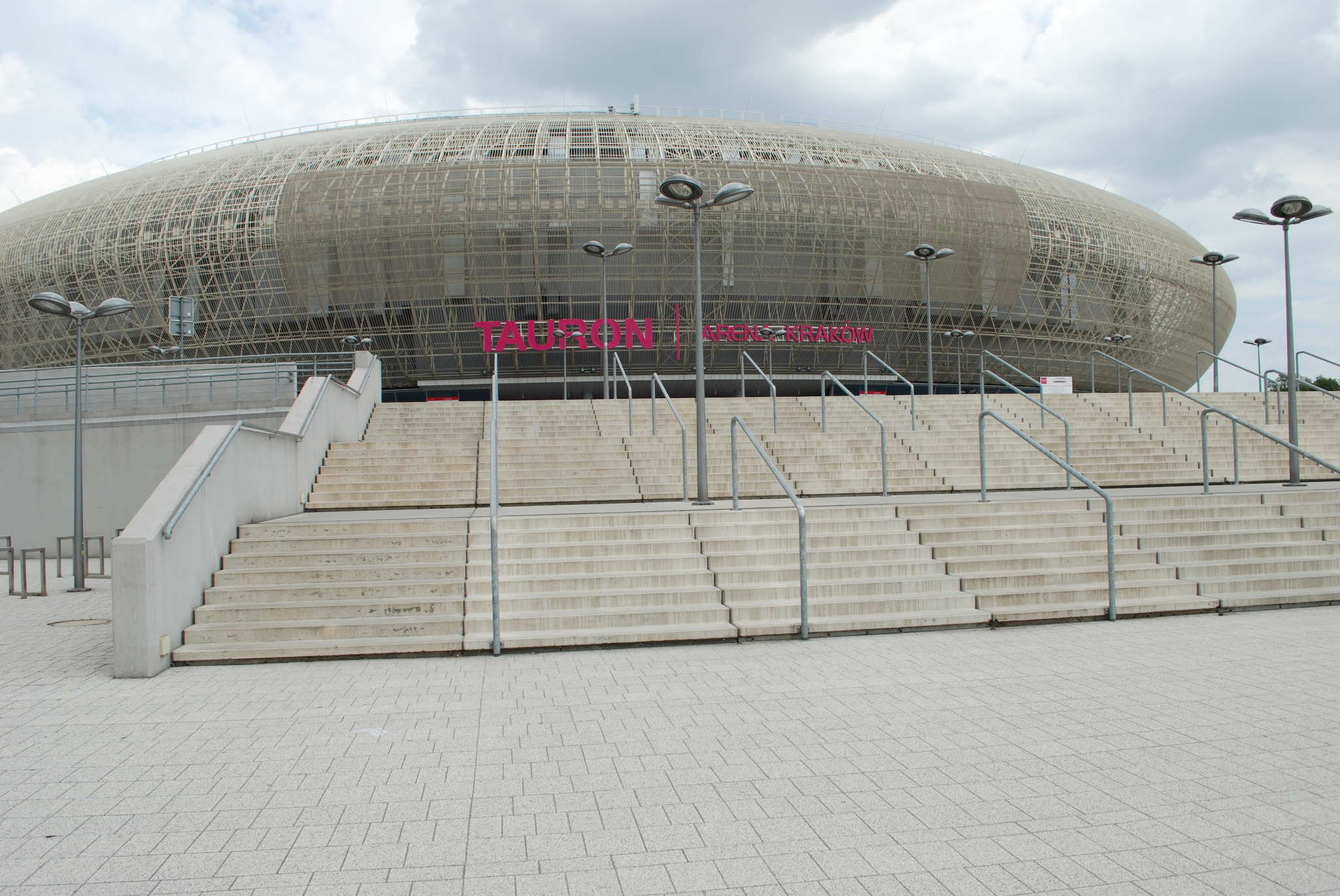 Szerokie, betonowe schody z metalowymi poręczami prowadzące do nowoczesnego stadionu z siatkową konstrukcją elewacji i dużym, różowym napisem na fasadzie.