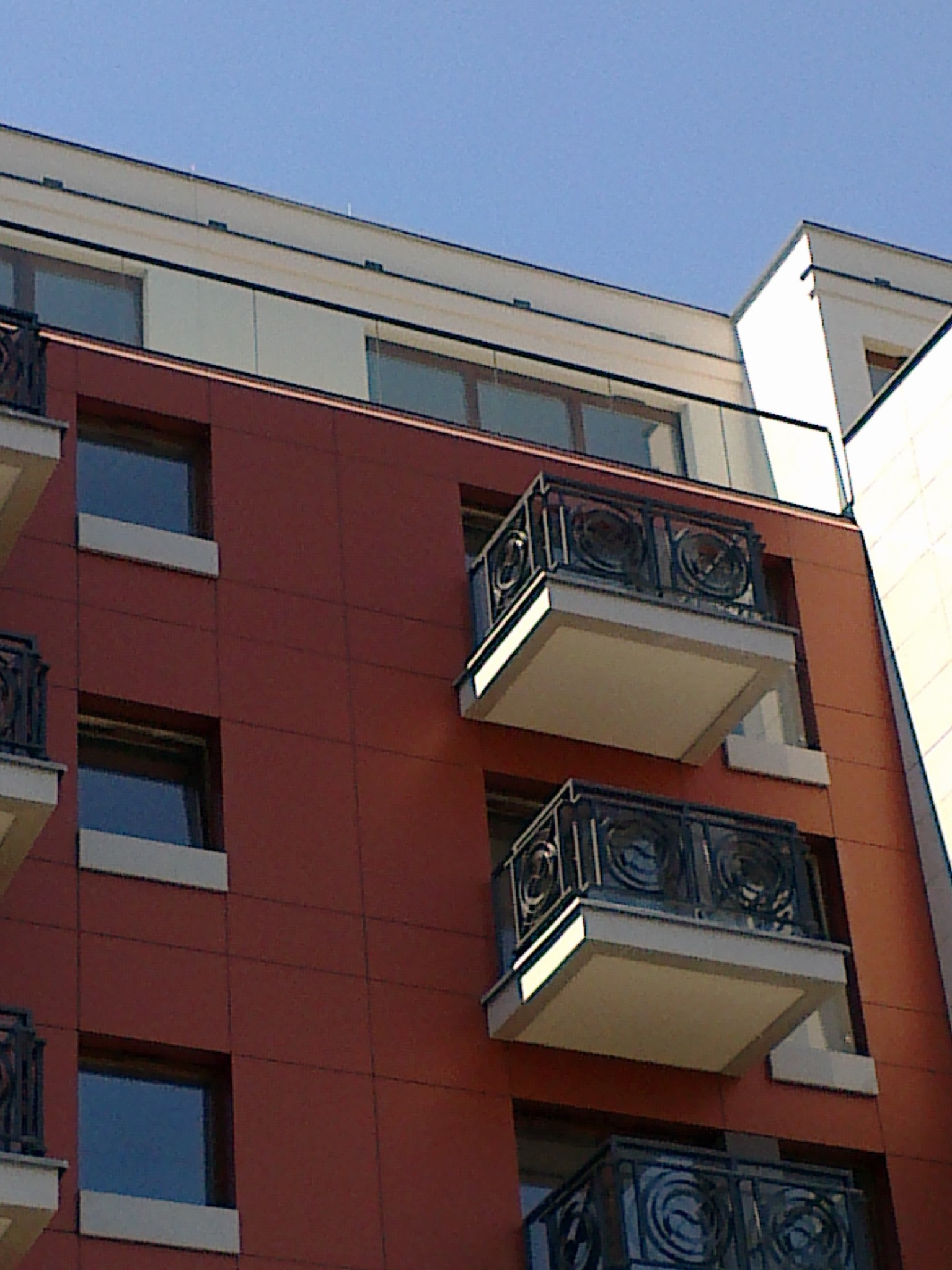 Widok z dołu na elewację budynku z balkonami z metalowymi balustradami o dekoracyjnym wzorze i szklanymi panelami, na tle błękitnego nieba.