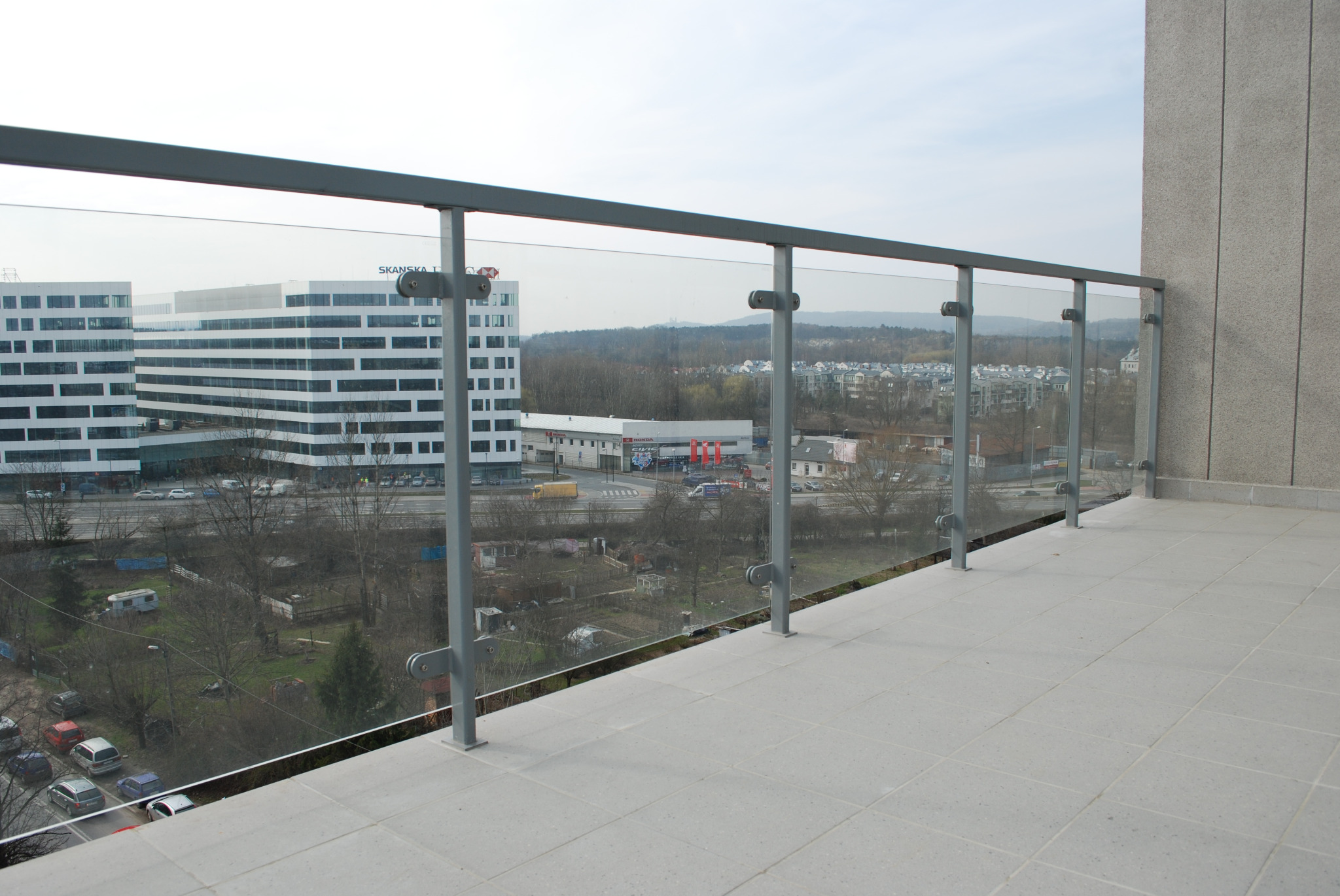 Balkon z szarą, metalową balustradą i szklanym wypełnieniem, widok na budynki biurowe i okolicę.