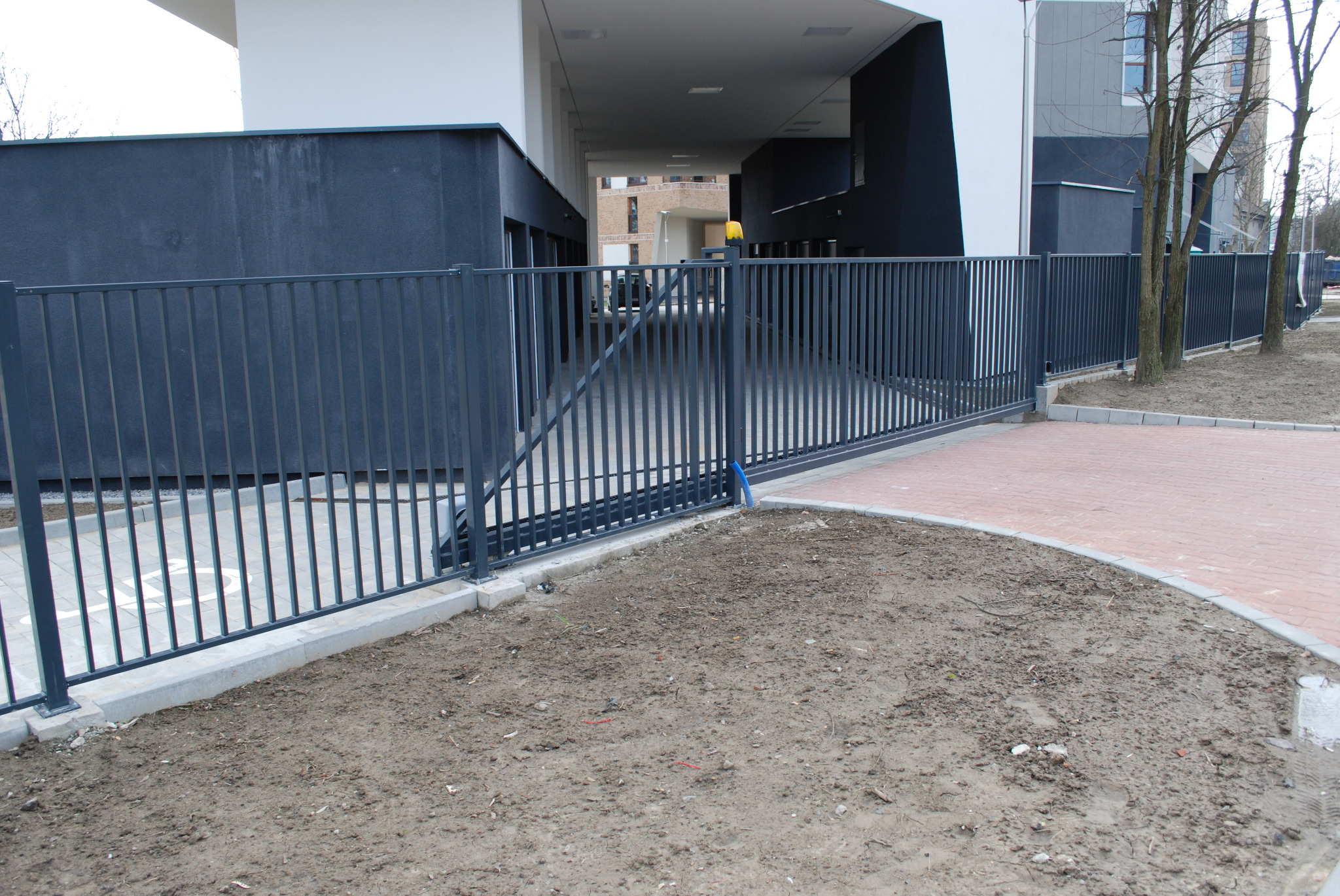 Nowoczesna, przesuwna brama z metalowymi prętami w kolorze grafitowym, zamontowana przed budynkiem mieszkalnym z widocznym napędem i sygnalizacją świetlną.