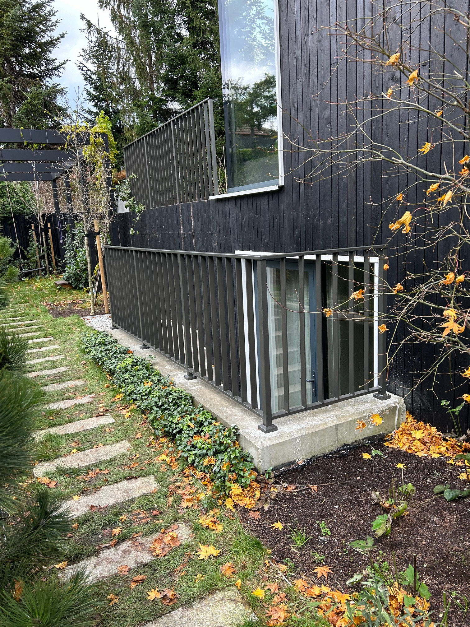 Ciemna, minimalistyczna balustrada metalowa przy oknie piwnicznym, otoczona zielenią i opadłymi liśćmi, z betonową ścieżką w tle.