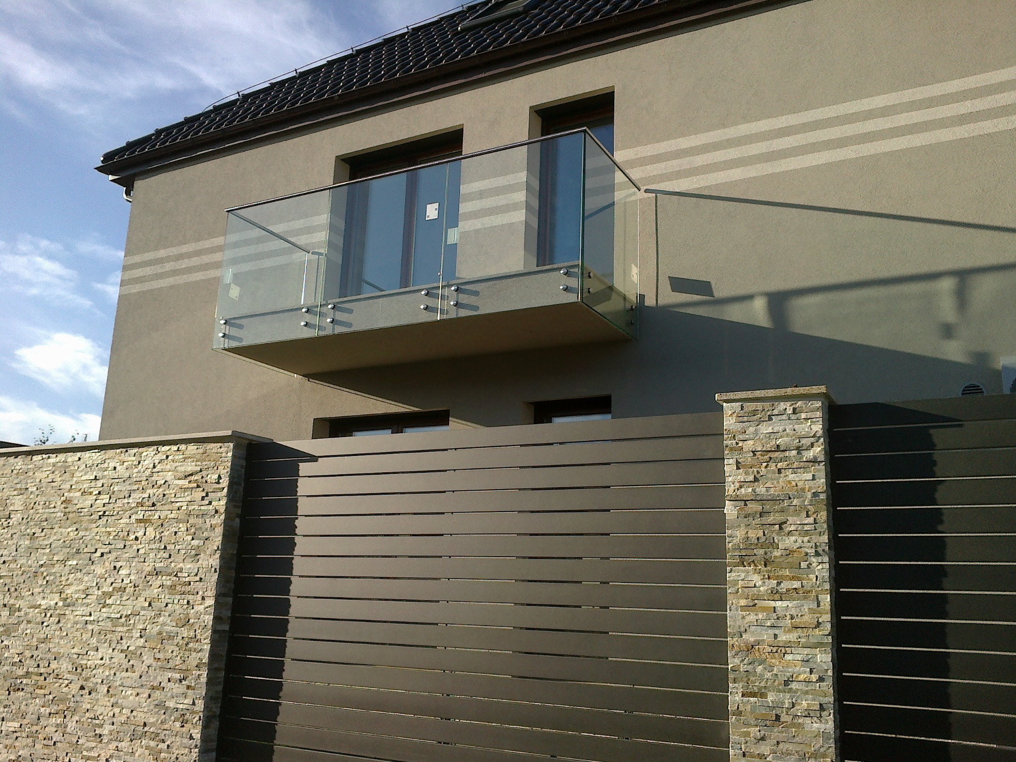 Nowoczesny balkon z szklaną balustradą mocowaną na metalowych wspornikach, widok z dołu na tle domu z szarą elewacją i metalowym ogrodzeniem z elementami kamiennymi.