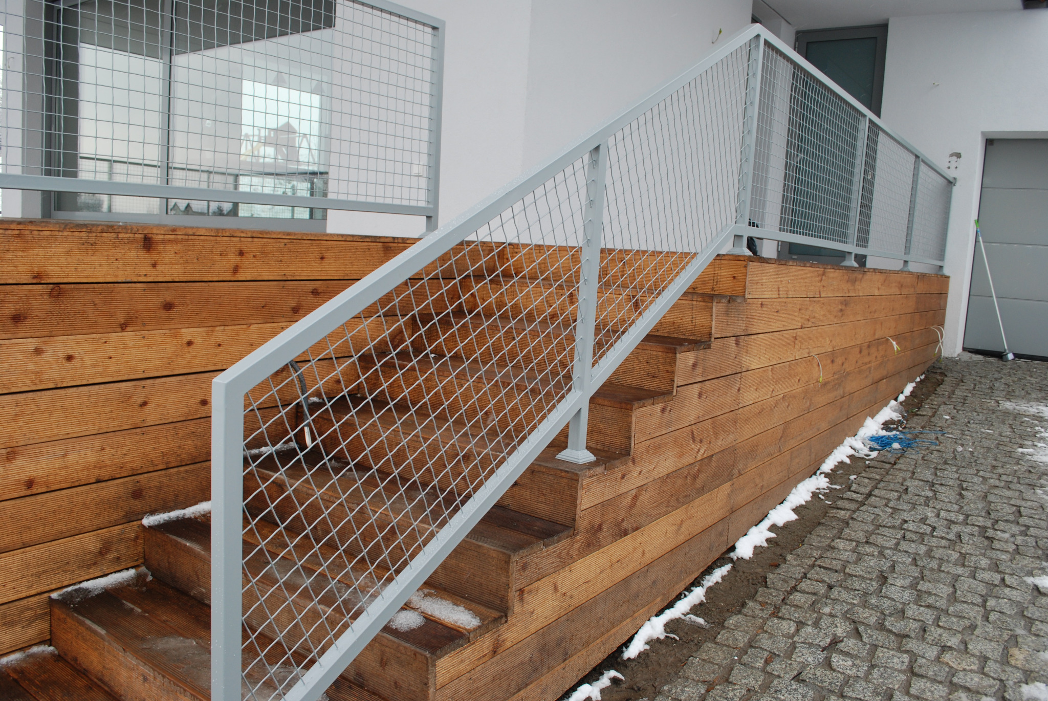 Zewnętrzne drewniane schody z siatkową balustradą w kolorze jasnoszarym, prowadzące do budynku z szarą bramą garażową i drewnianą elewacją, na ziemi resztki śniegu.