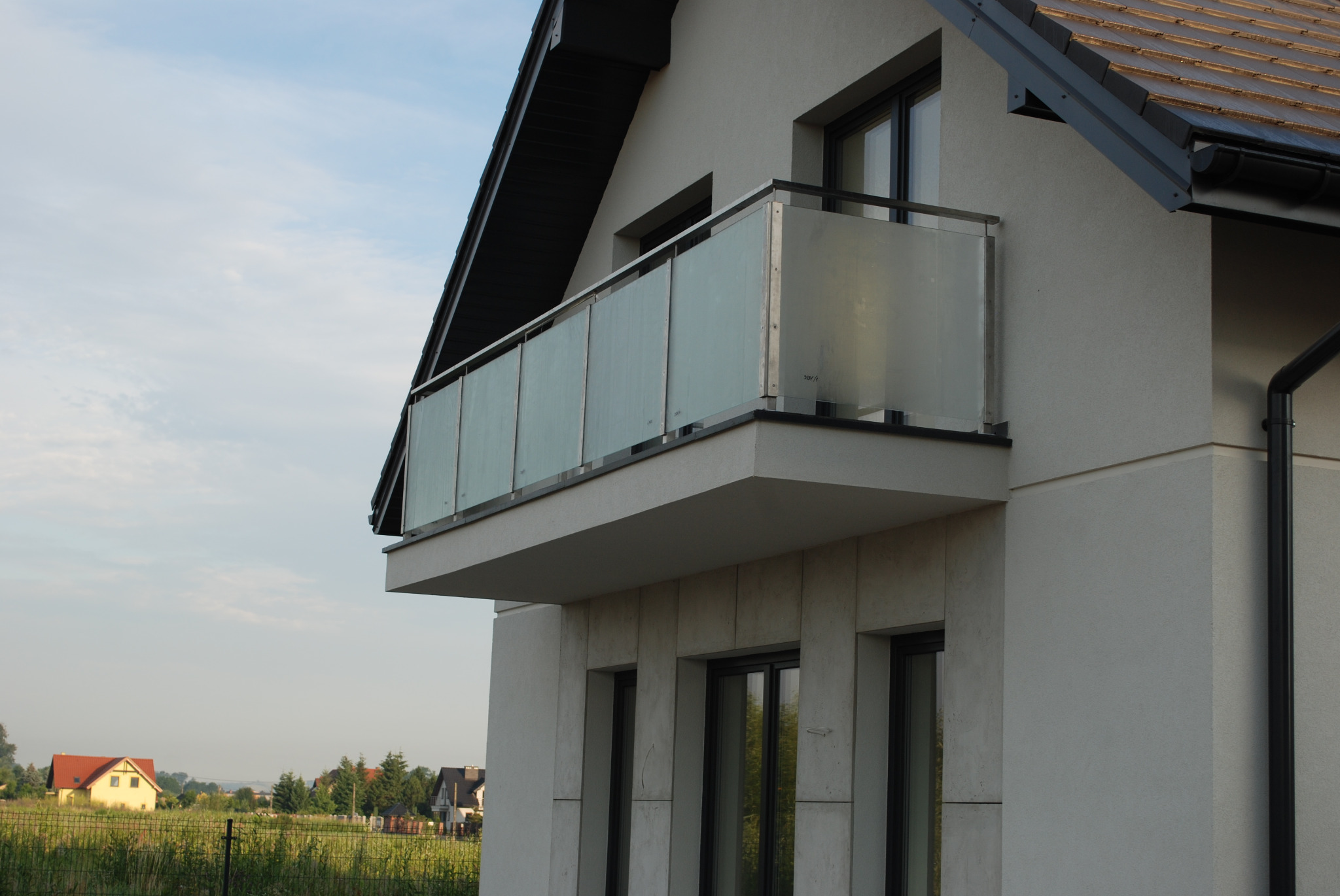 Nowoczesny balkon z szklaną balustradą na tle jasnego budynku z ciemnym dachem i widokiem na okoliczne domy i pola.