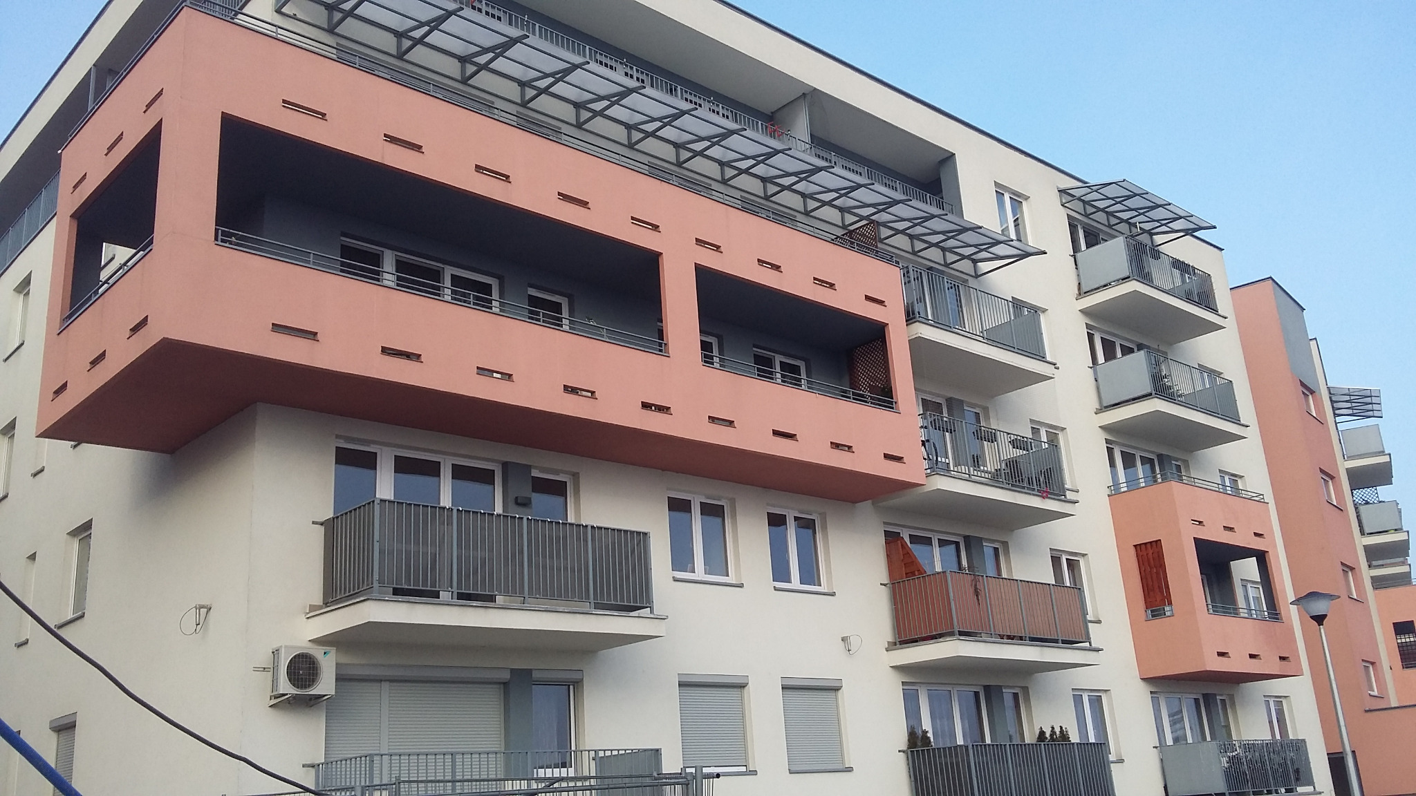 Nowoczesny budynek mieszkalny z balkonami zabezpieczonymi metalowymi barierkami, widoczne elementy konstrukcji zadaszenia nad balkonami na wyższych piętrach.