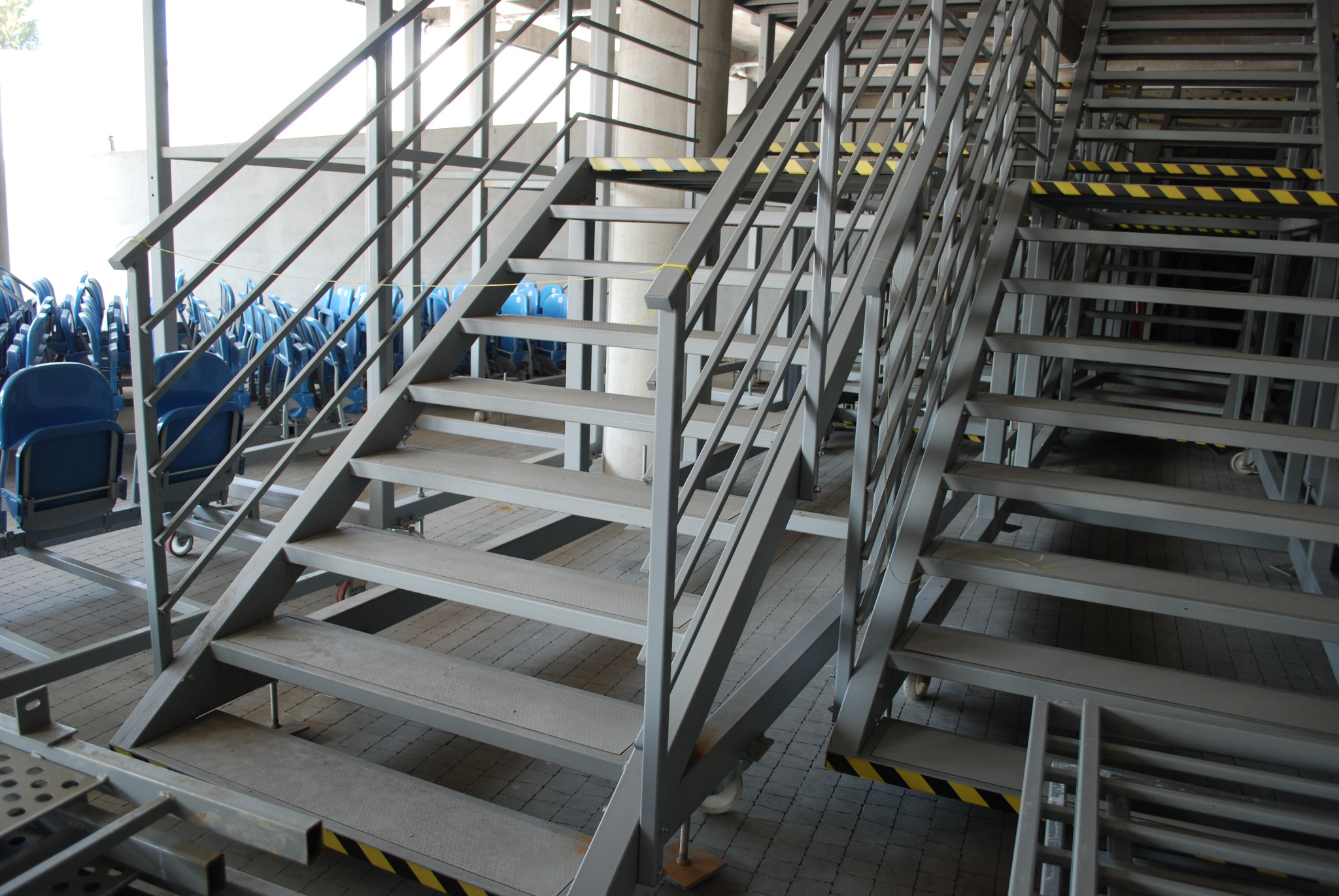 Metalowe schody z szarą konstrukcją i żółto-czarnymi pasami ostrzegawczymi, widoczne krzesła na kółkach w tle.