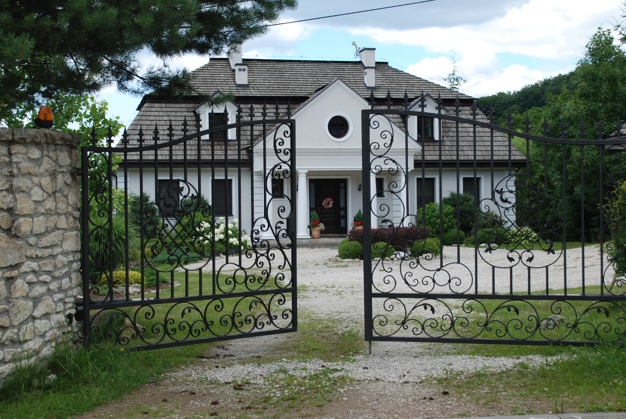Otwarta, czarna, ozdobna brama metalowa z dekoracyjnymi elementami na tle białego domu z szarą dachówką i kamiennego ogrodzenia.