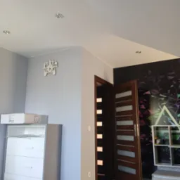 Pokój po remoncie: widok na drewniane drzwi z przeszkleniami, komodę z szufladami, dekoracyjny zegar w kształcie pada do gier na szarej ścianie oraz wzorzystą tapetę z motywem geometrycznym.