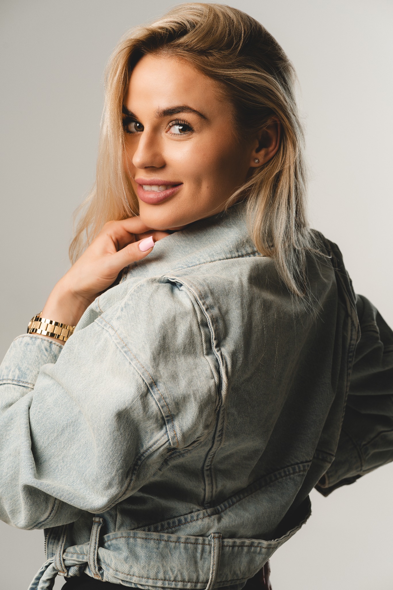 Uśmiechnięta blondynka w jeansowej kurtce, spoglądająca przez ramię, z widocznym złotym zegarkiem na nadgarstku i delikatnym makijażem.