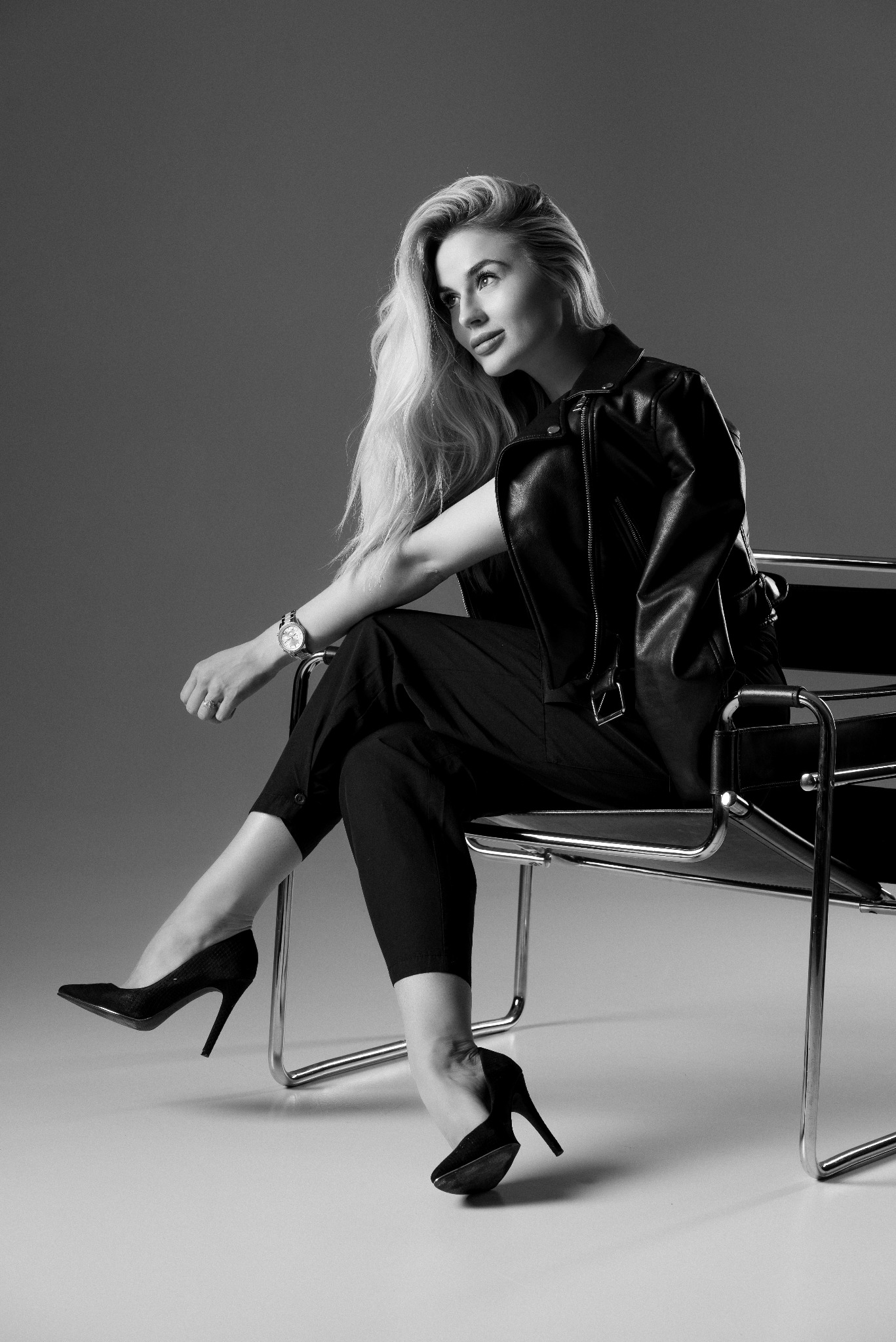 Czarno-białe zdjęcie kobiety w czarnym skórzanym żakiecie i spodniach, siedzącej na chromowanym krześle Marcela Breuera z zegarkiem na ręce i czarnych szpilkach, spoglądającej w lewo.
