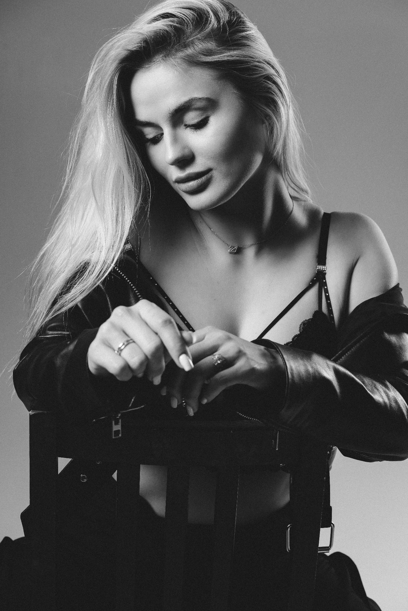 Czarno-biały portret kobiety z długimi blond włosami, w skórzanej kurtce i delikatnej biżuterii, siedzącej na krześle z zamkniętymi oczami i splecionymi dłońmi