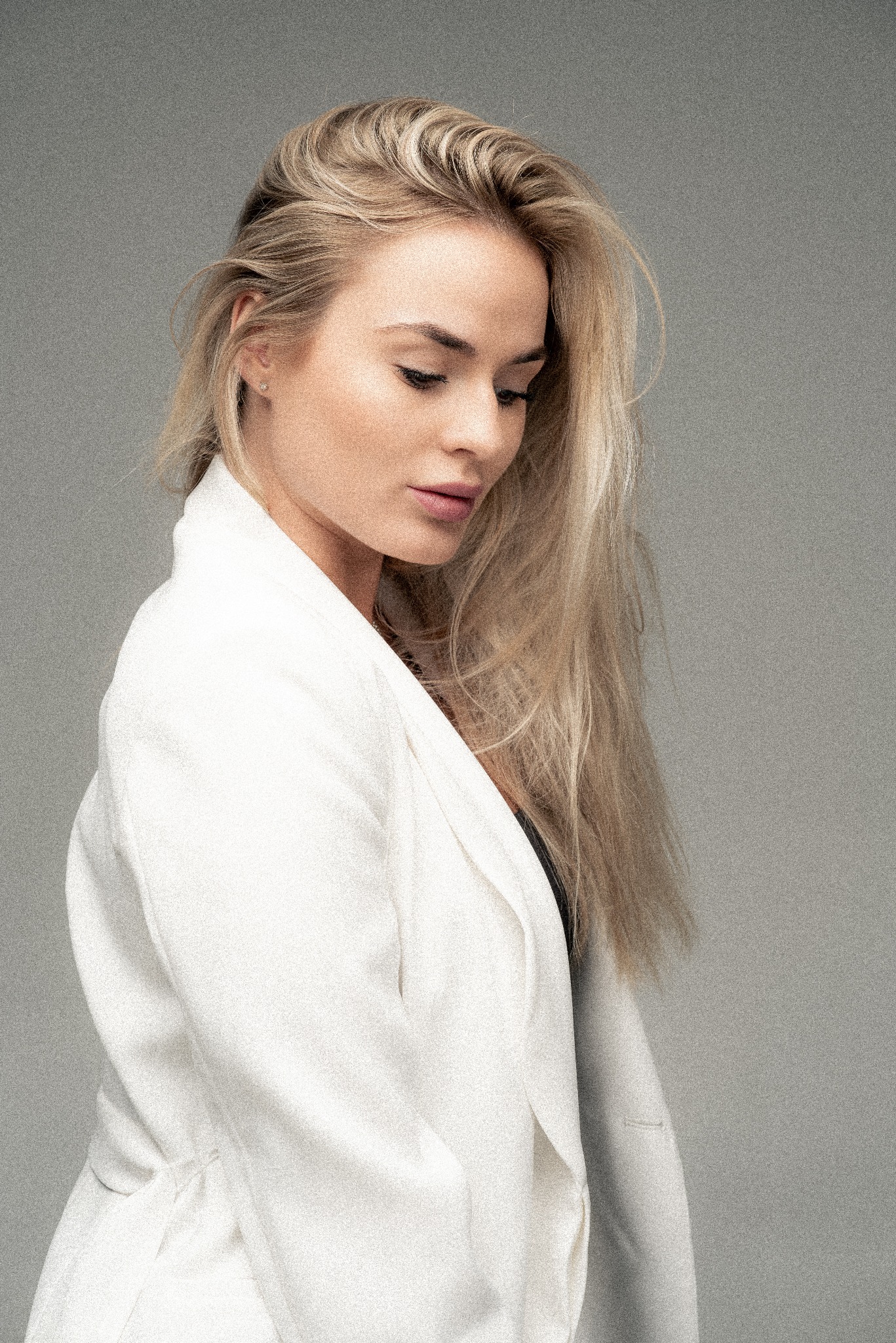 Portret kobiety z blond włosami w białym żakiecie na szarym tle, spuszczony wzrok, subtelny makijaż.