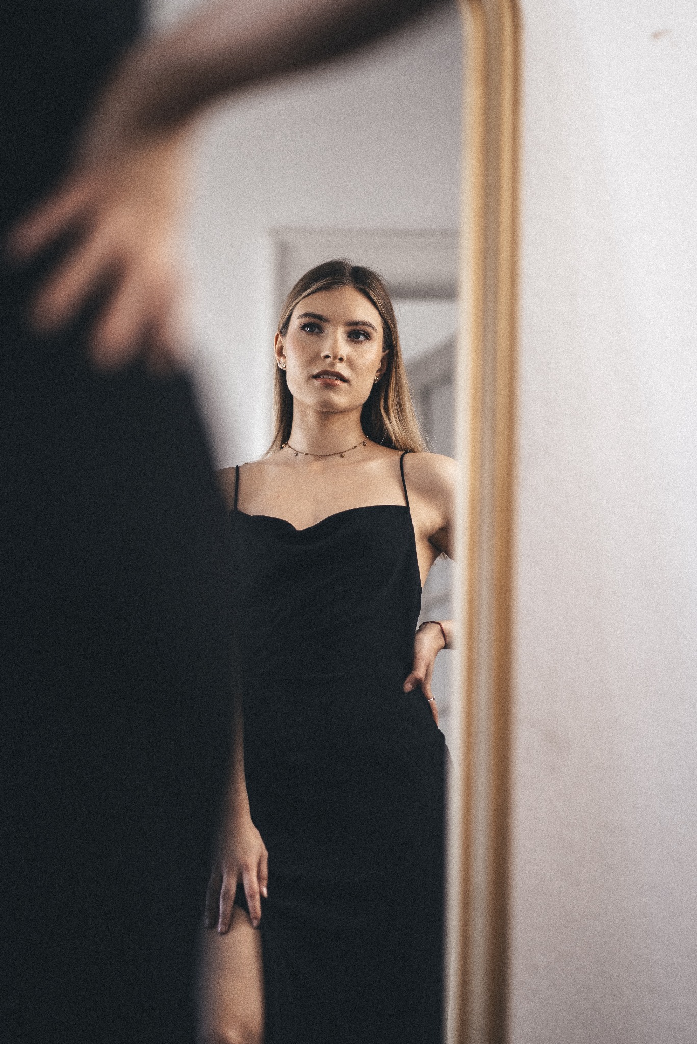 Młoda kobieta w czarnej sukience na ramiączkach, pozująca przed lustrem w złotej ramie, z widocznym odbiciem sylwetki w tle.