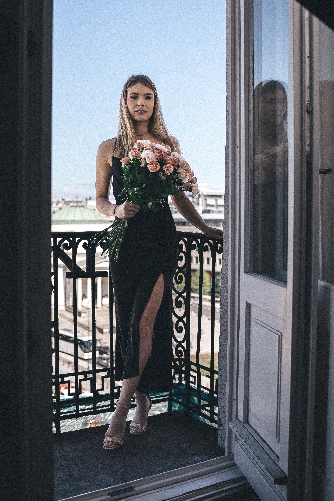 Młoda kobieta w czarnej sukni z rozcięciem, trzymająca bukiet różowych róż, pozuje na balkonie z ozdobną balustradą, z widokiem na miasto w tle.