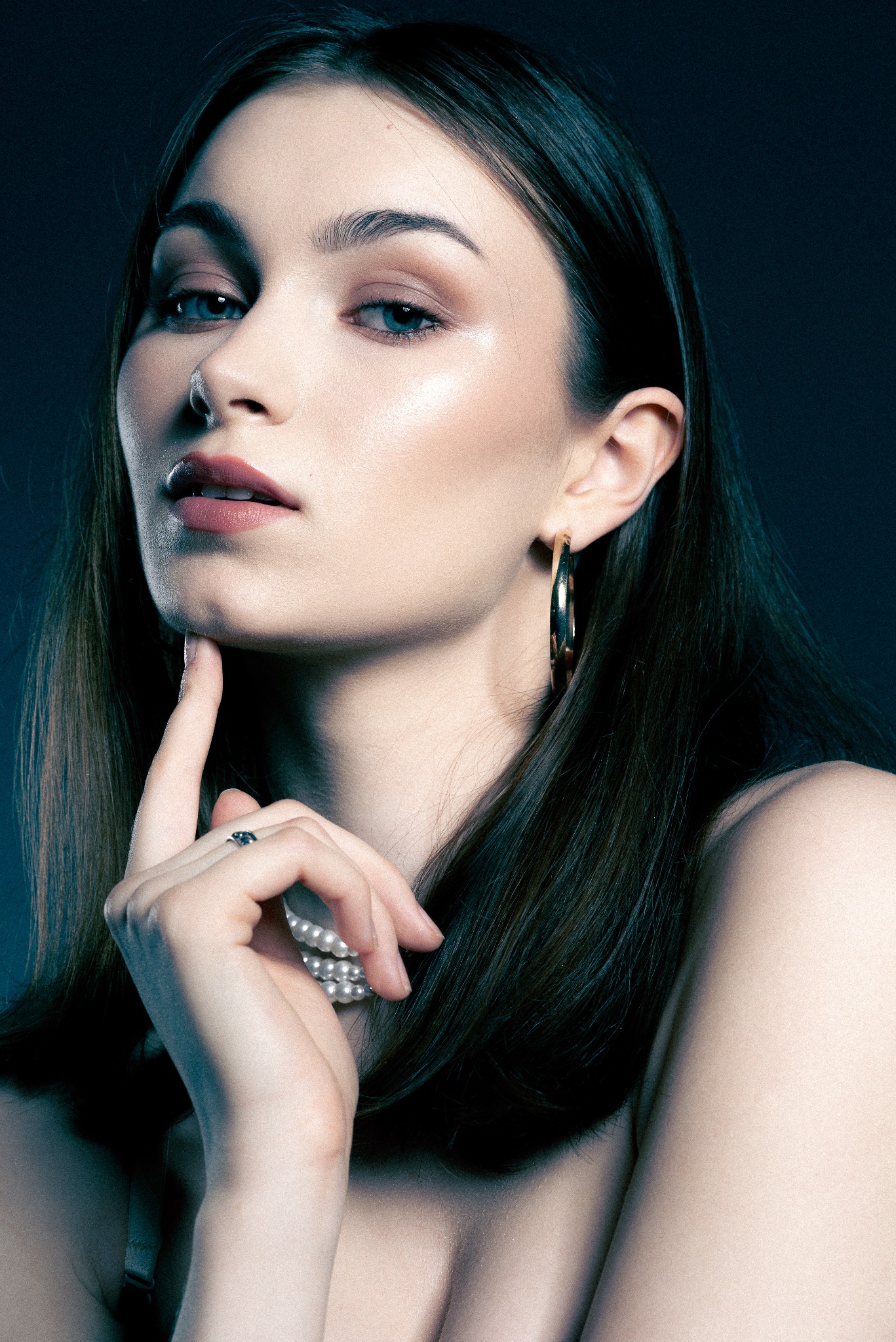 Portret kobiety z platynowymi kolczykami kołami, perłami i pierścionkiem z ciemnym oczkiem, dotykającej palcem brody na ciemnym tle.