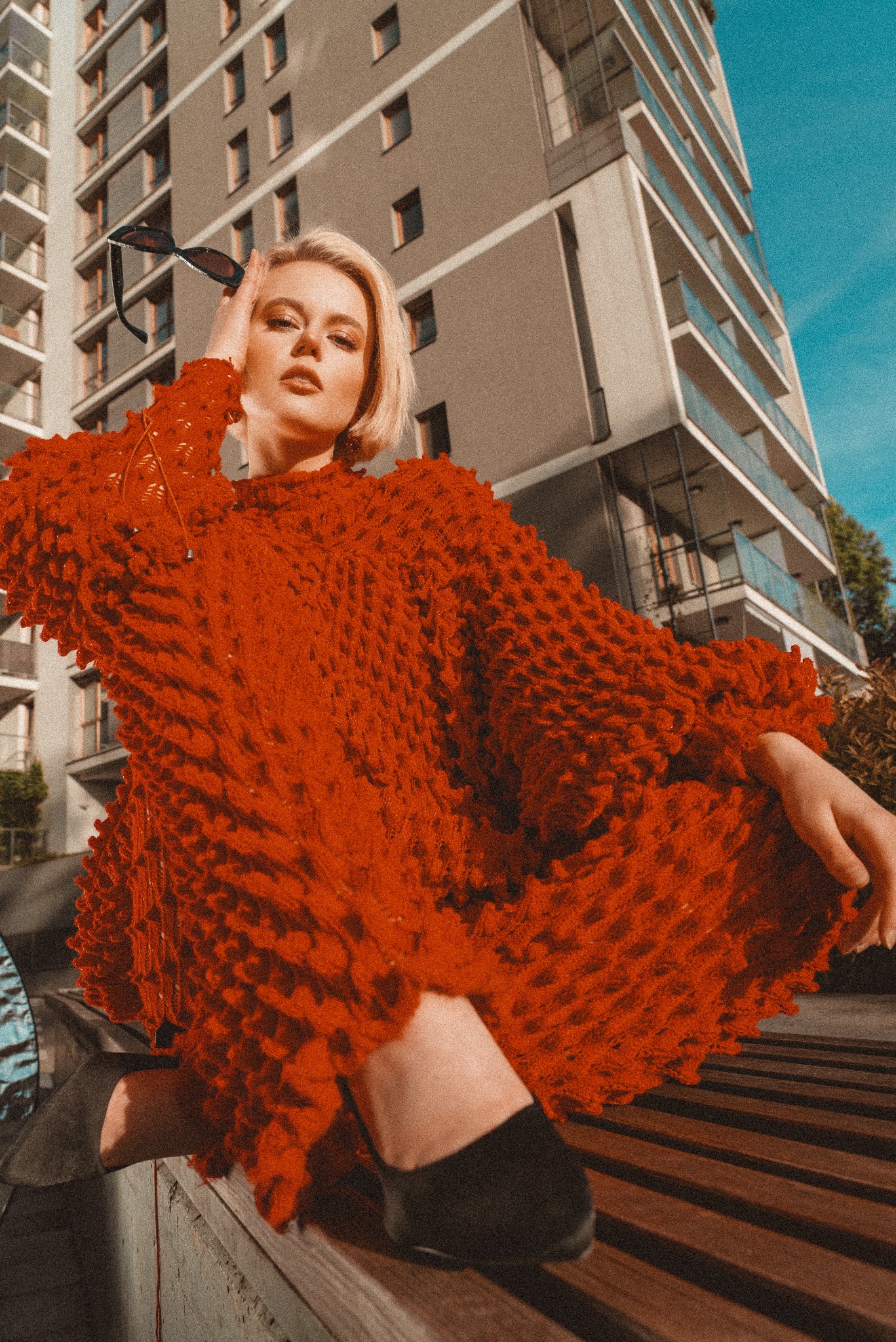 Kobieta z blond włosami w czerwonym, teksturowanym swetrze, pozuje na tle nowoczesnego budynku, trzymając okulary przeciwsłoneczne przy głowie.