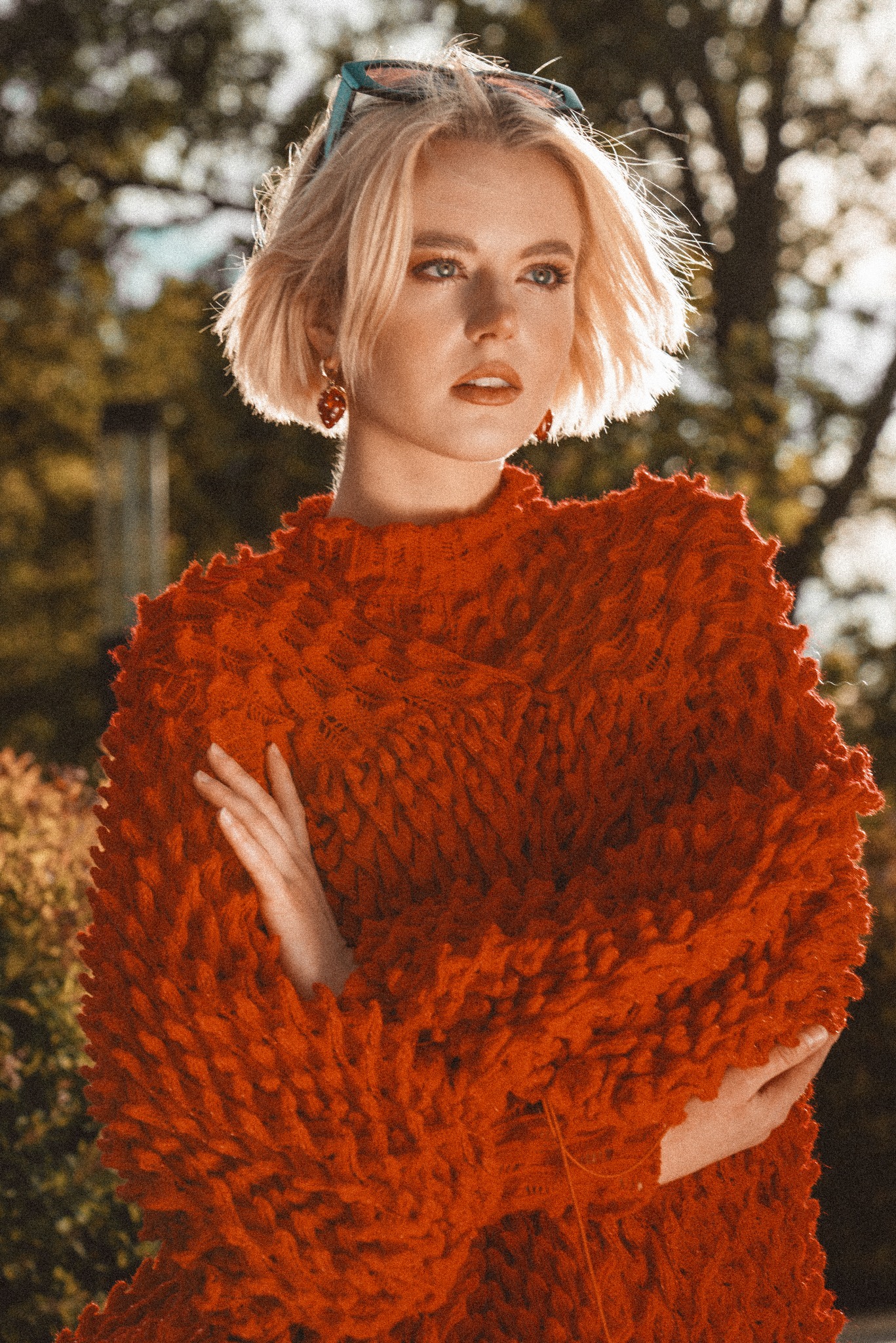 Portret kobiety o krótkich blond włosach w pomarańczowym, teksturowanym swetrze, z okularami przeciwsłonecznymi na głowie i kolczykami w kształcie owoców, pozującej na tle drzew