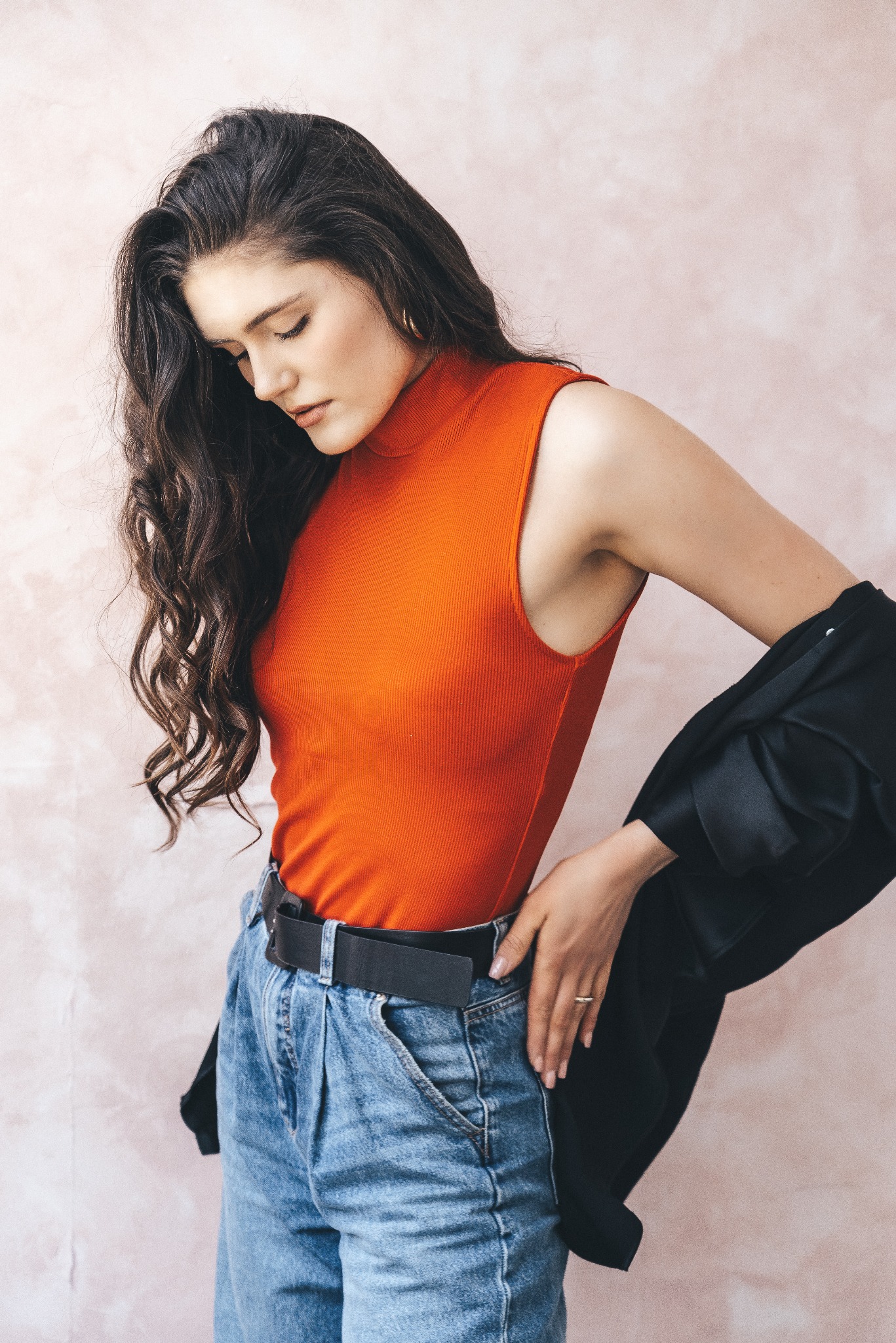 Portret kobiety w pomarańczowym golfie i jeansach, z czarną marynarką nonszalancko zarzuconą na ramię, pozującej na tle delikatnej, różowej ściany.