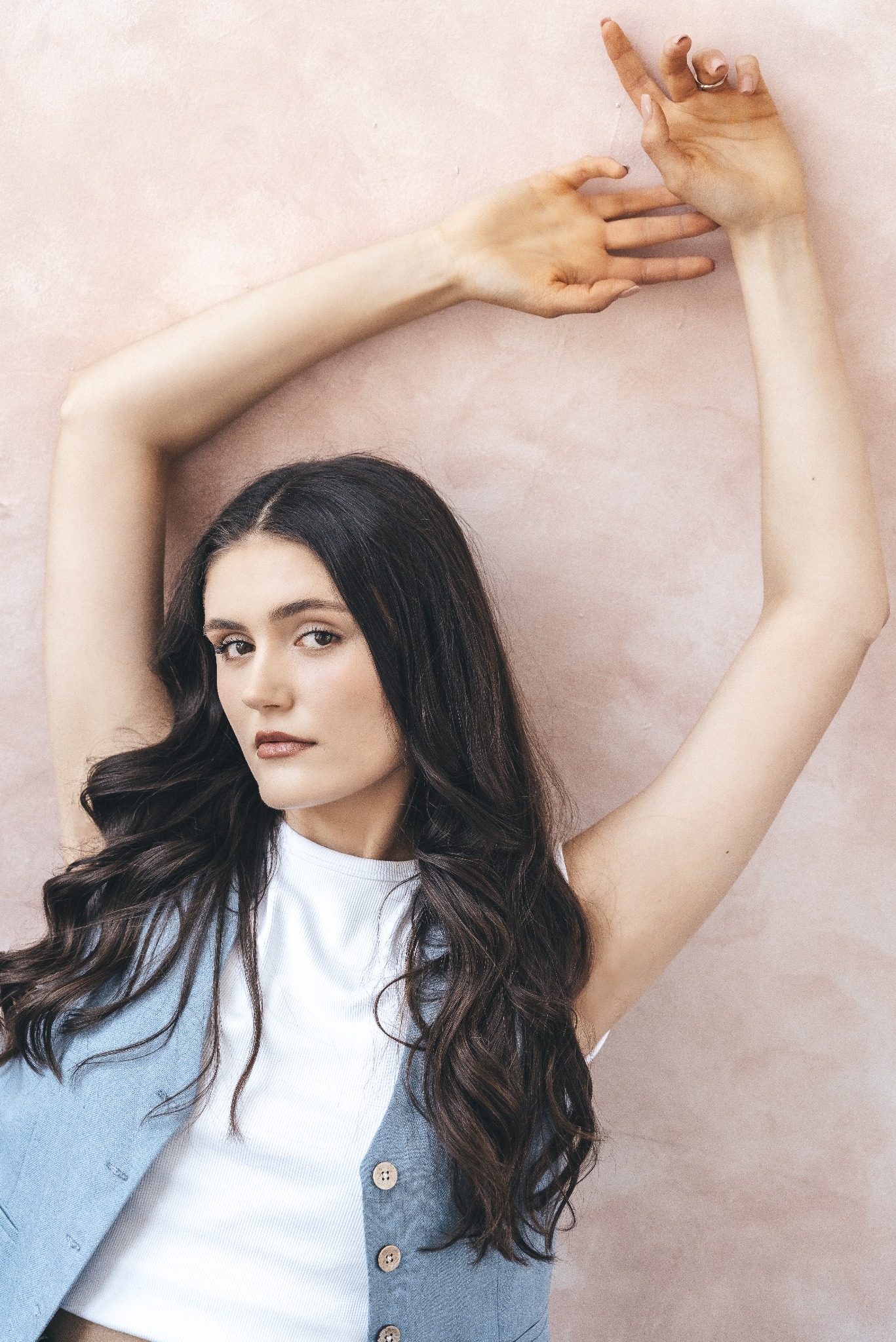 Portret kobiety z ciemnymi, falowanymi włosami, ubranej w białą koszulkę i dżinsową kamizelkę, pozującej z uniesionymi rękami na tle teksturowanej, różowej ściany.