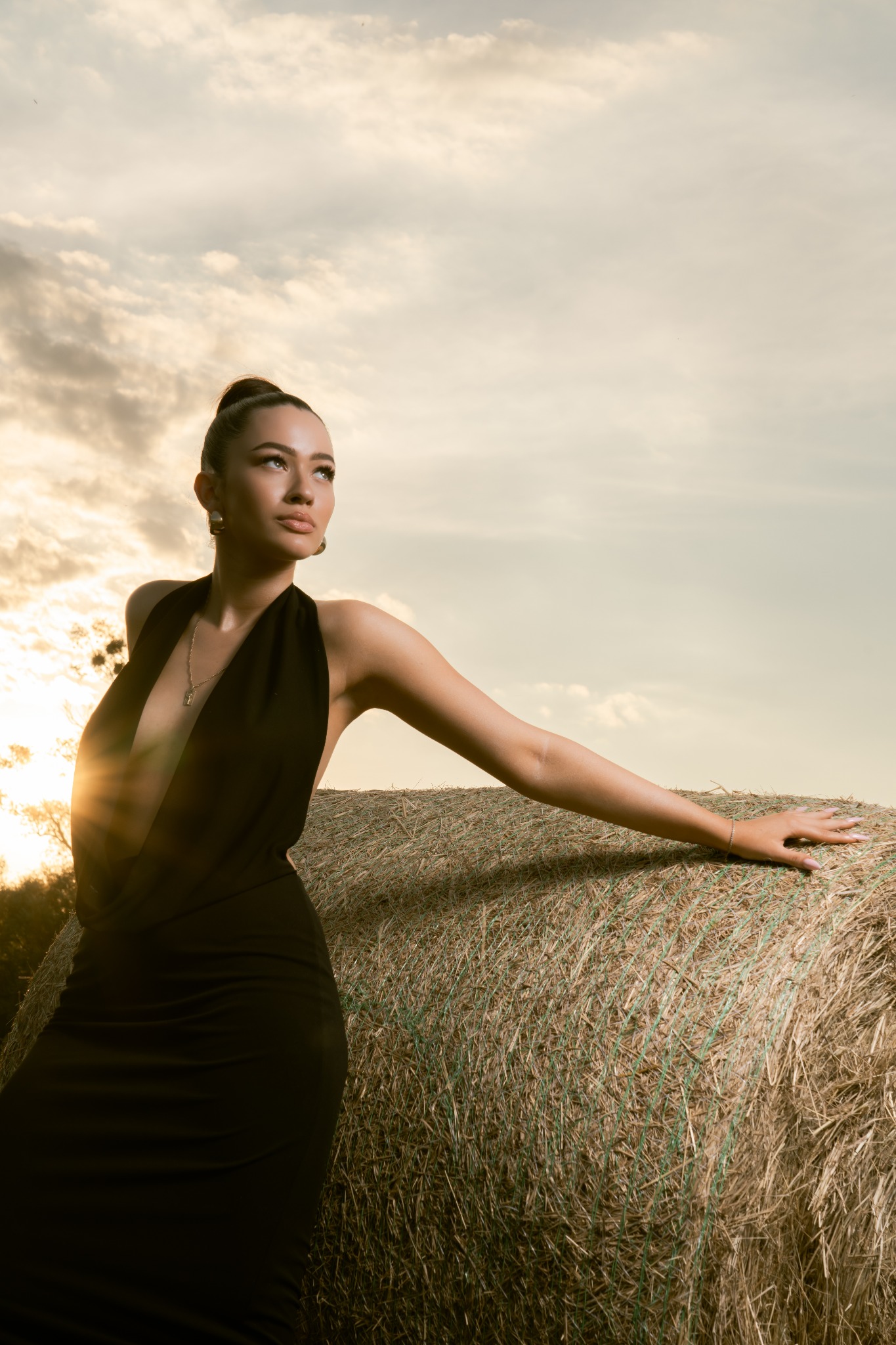Kobieta w czarnej sukni pozuje opierając się o belę siana na tle zachodzącego słońca, z delikatnym uśmiechem i spojrzeniem skierowanym w górę.