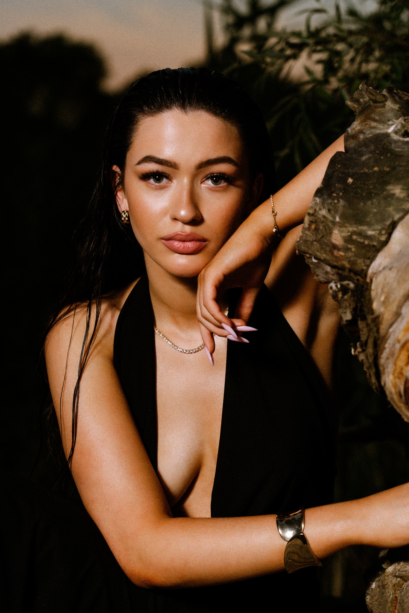 Portret kobiety w czarnej sukience z głębokim dekoltem, opierającej się o drewno, z widoczną biżuterią: kolczyki, naszyjnik i bransoletka.