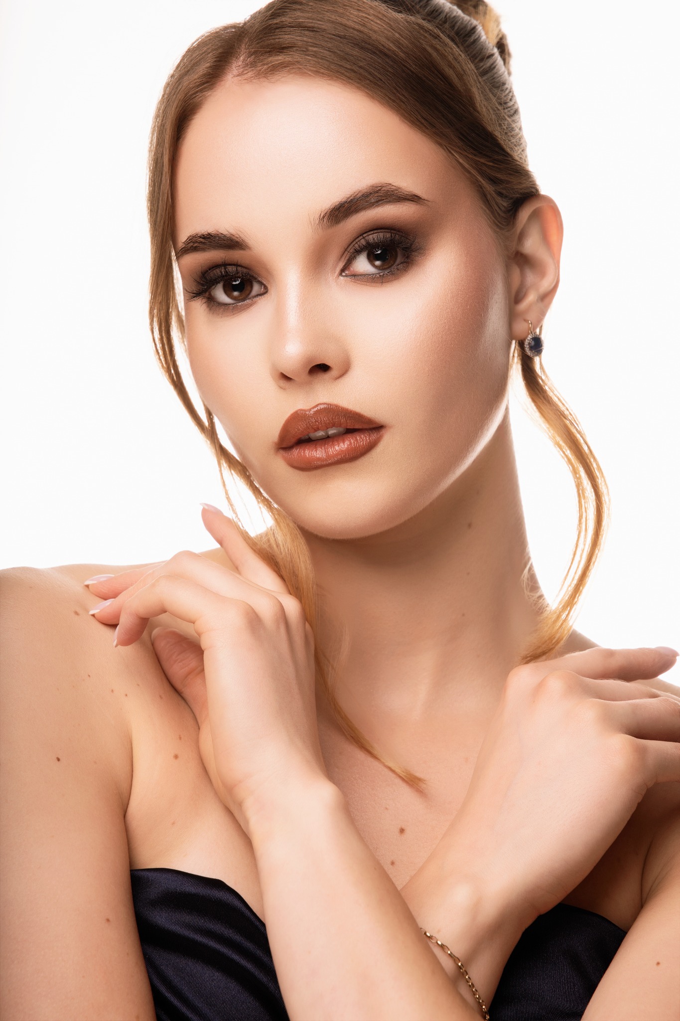 Portret młodej kobiety z delikatnym makijażem, błyszczącymi ustami i upiętymi włosami, pozującej na białym tle z dłońmi skrzyżowanymi na ramionach, widoczne kolczyki z ciemnymi kamieniami.