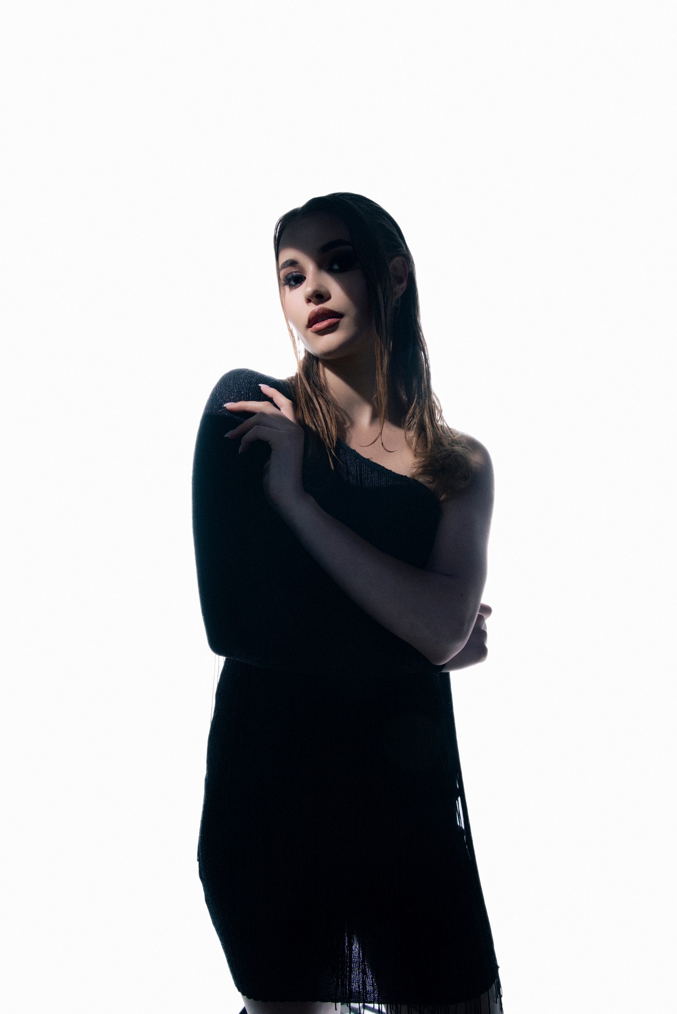 Portret kobiety w czarnej sukience na jednym ramieniu, z mokrymi włosami i mocnym makijażem oczu, oświetlonej z góry, tworząc dramatyczny efekt cieni.