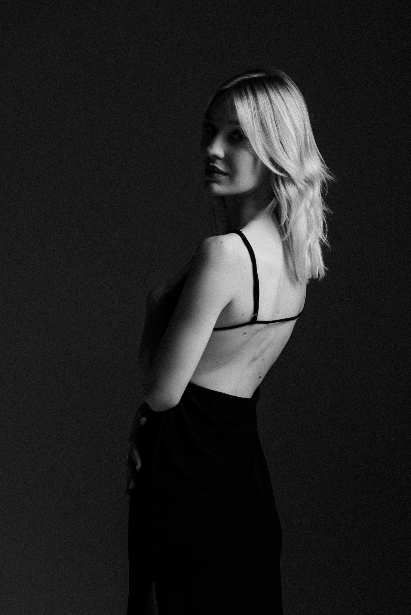 Czarno-białe zdjęcie kobiety z blond włosami, odwróconej przez ramię, w sukience na cienkich ramiączkach na ciemnym tle.