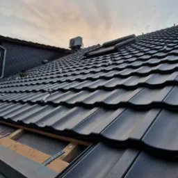 Częściowo pokryty dach z czarnej dachówki ceramicznej, widoczna drewniana konstrukcja łatowania i mocowanie do rynny, klimatyzator na dachu sąsiedniego budynku.