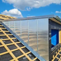 Konstrukcja dachu z lukarną obłożoną blachą, widoczne łaty, membrana dachowa z nadrukiem, błękitne niebo w tle.