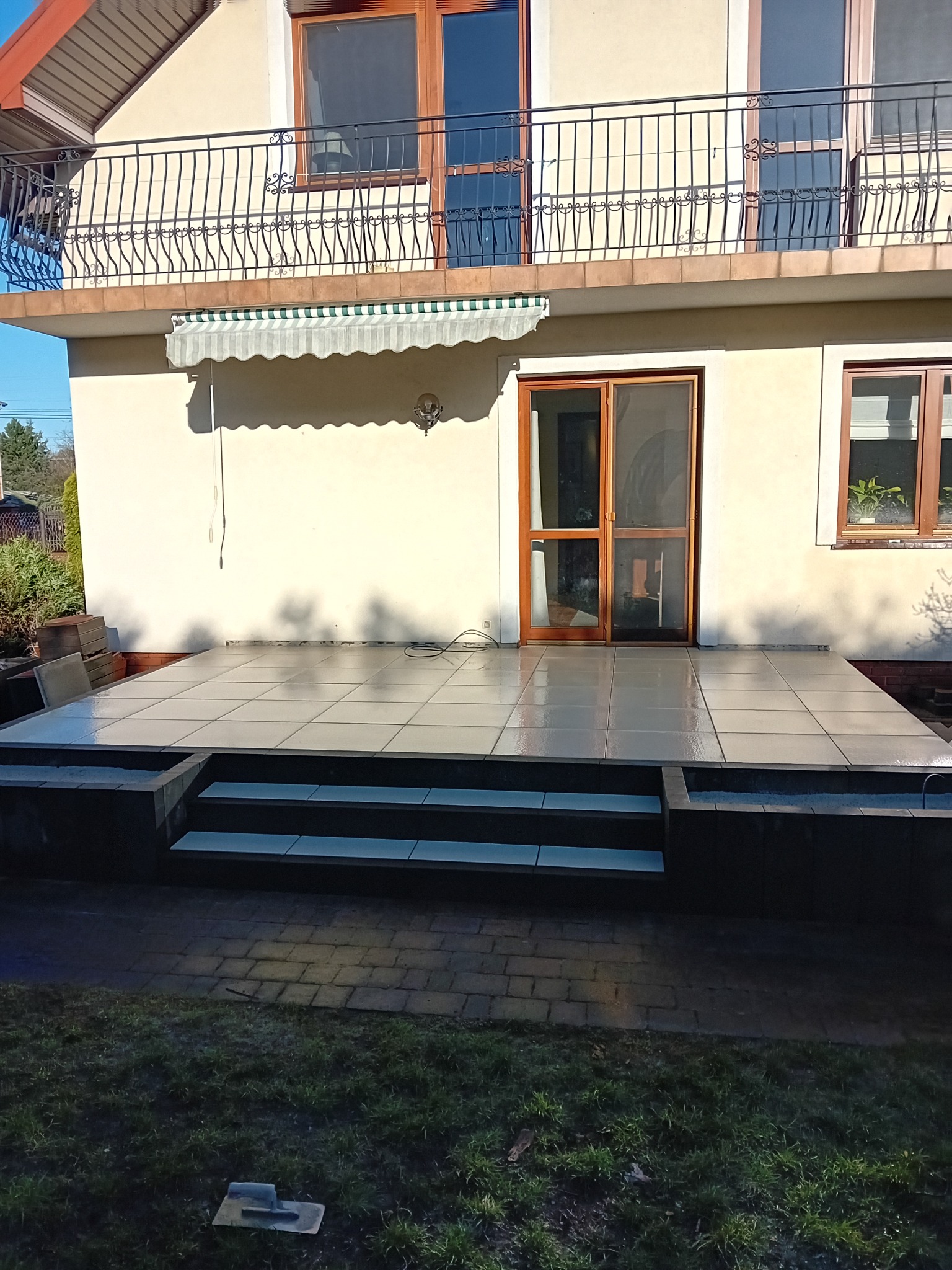 Nowo wyłożony, jasnoszary taras z płytek ceramicznych z trzema stopniami, prowadzący do drzwi balkonowych domu z beżową elewacją i czarną balustradą balkonową na piętrze.