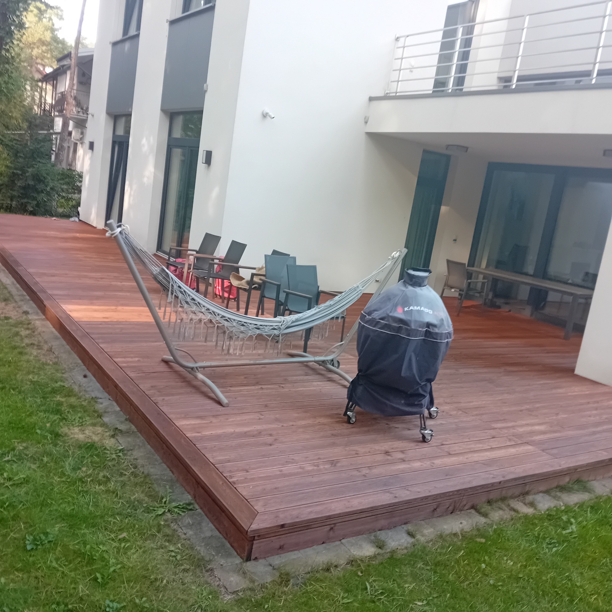 Nowoczesny taras z drewna z hamakiem i grillem Kamado, przylegający do białego budynku z dużymi oknami, otoczony trawnikiem.