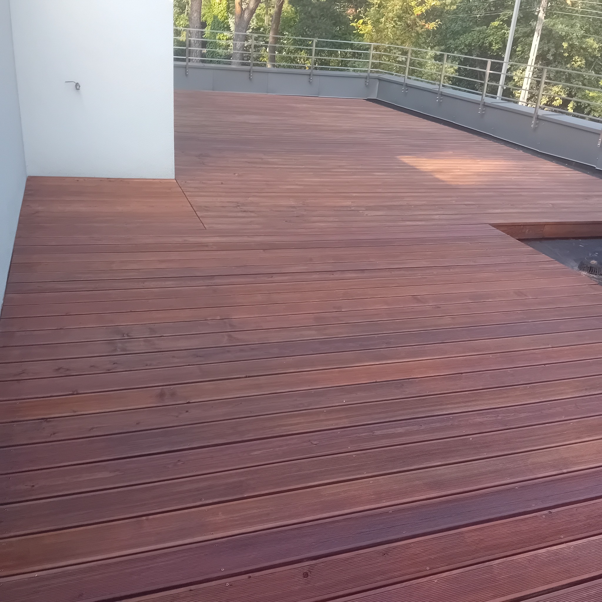 Rozległy taras z desek o ciepłym, brązowym odcieniu, z metalową balustradą w tle i fragmentem białej ściany po lewej stronie, ukazujący minimalistyczny design i precyzyjne wykonanie.