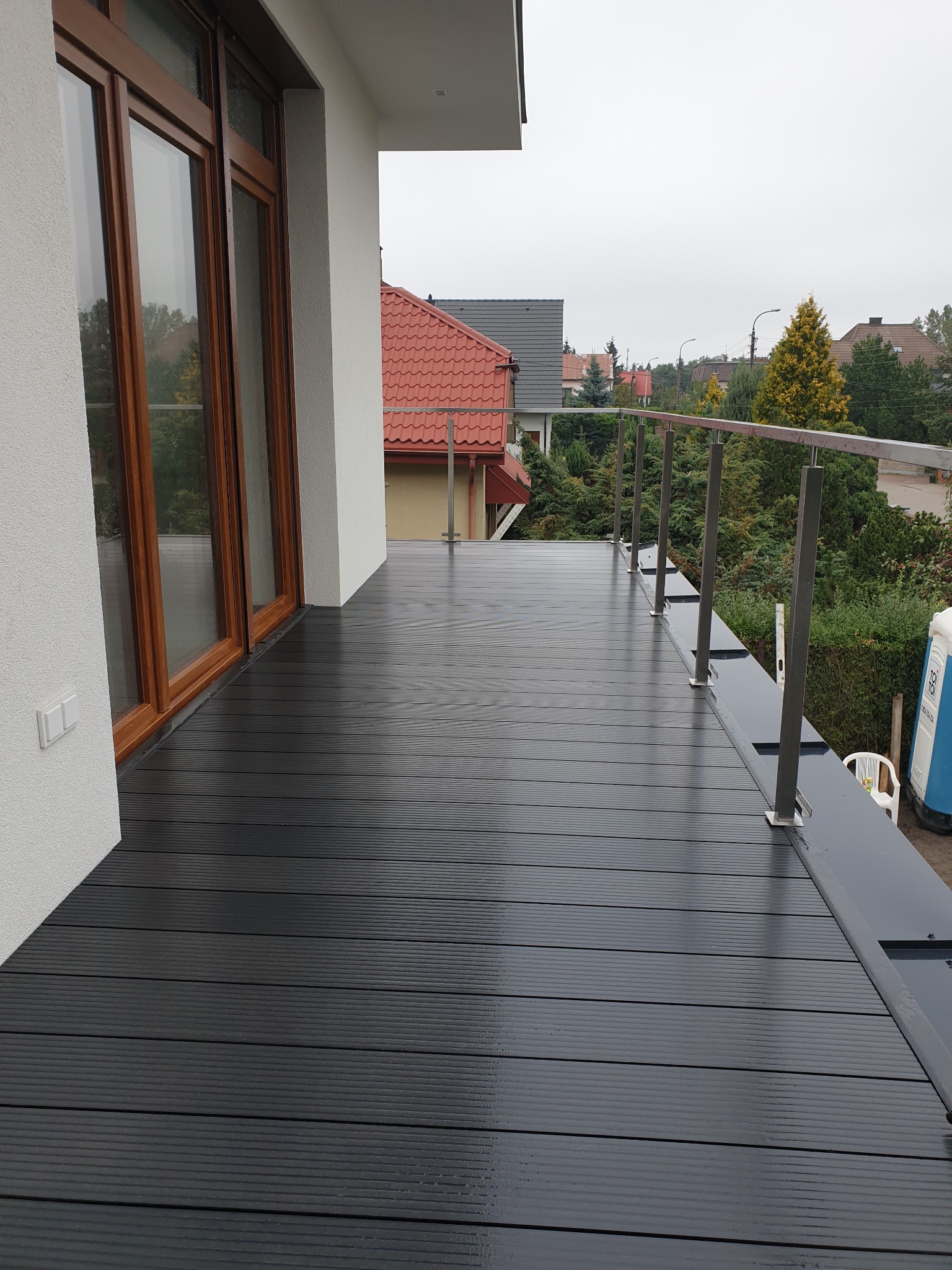 Nowoczesny taras z czarnej deski kompozytowej z metalową balustradą, widok na okoliczne domy w pochmurny dzień.