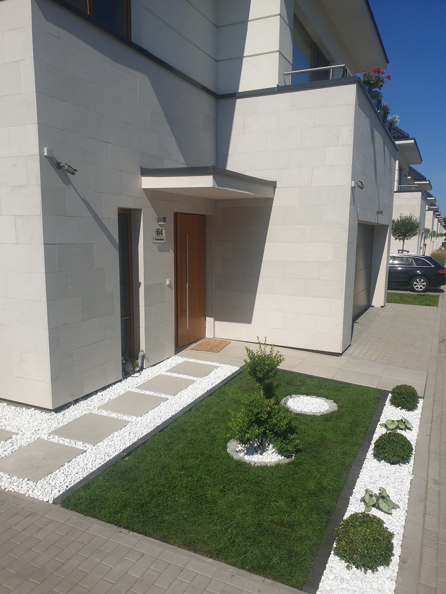 Nowoczesny ogród przed domem z trawnikiem, kamieniami dekoracyjnymi i ścieżką z płyt betonowych prowadzącą do drewnianych drzwi wejściowych.