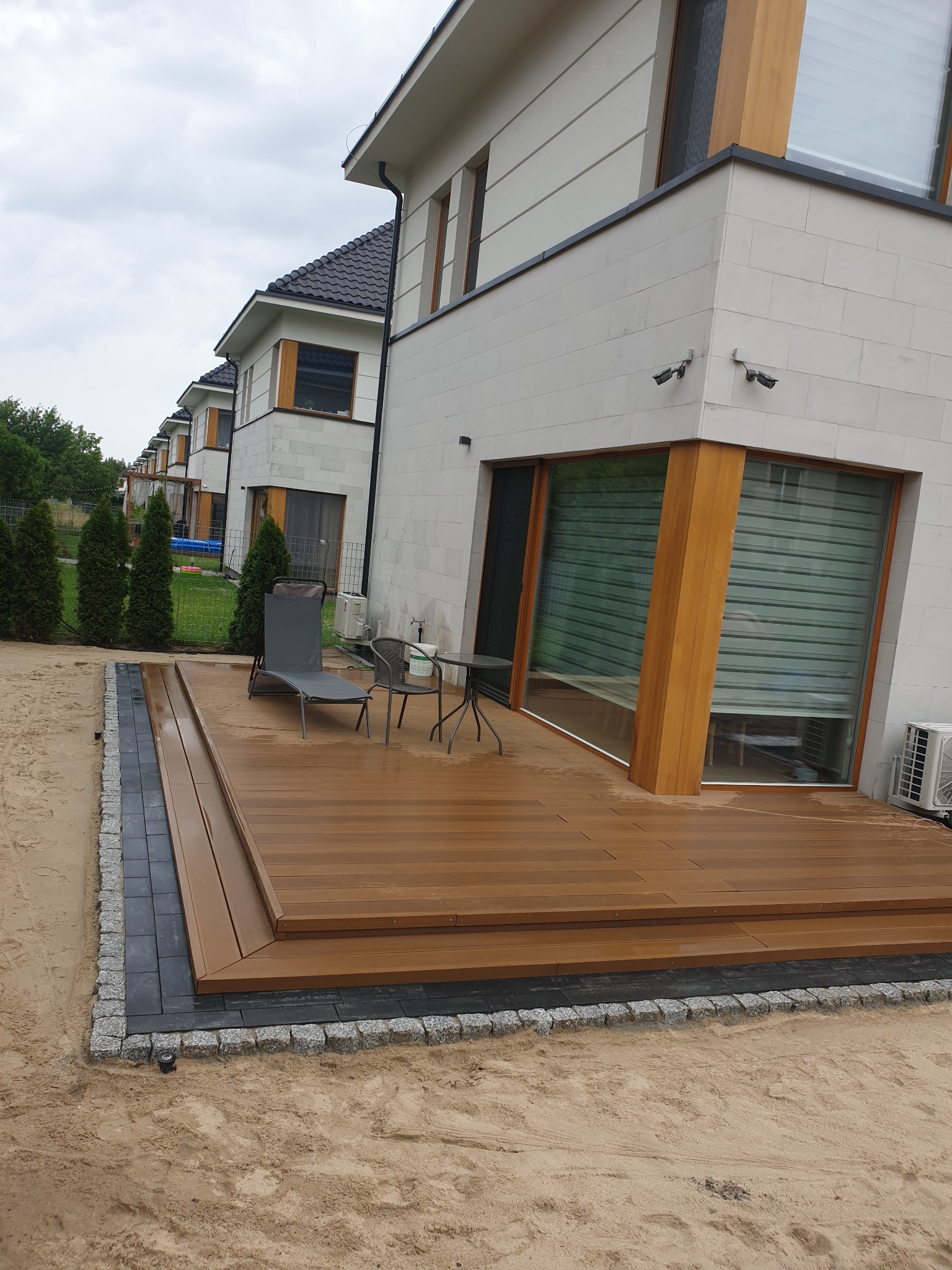 Nowoczesny taras z desek kompozytowych, obrzeża z kostki granitowej, szary leżak i stolik kawowy, tło stanowi ściana budynku z dużym oknem i szereg identycznych domów w tle.