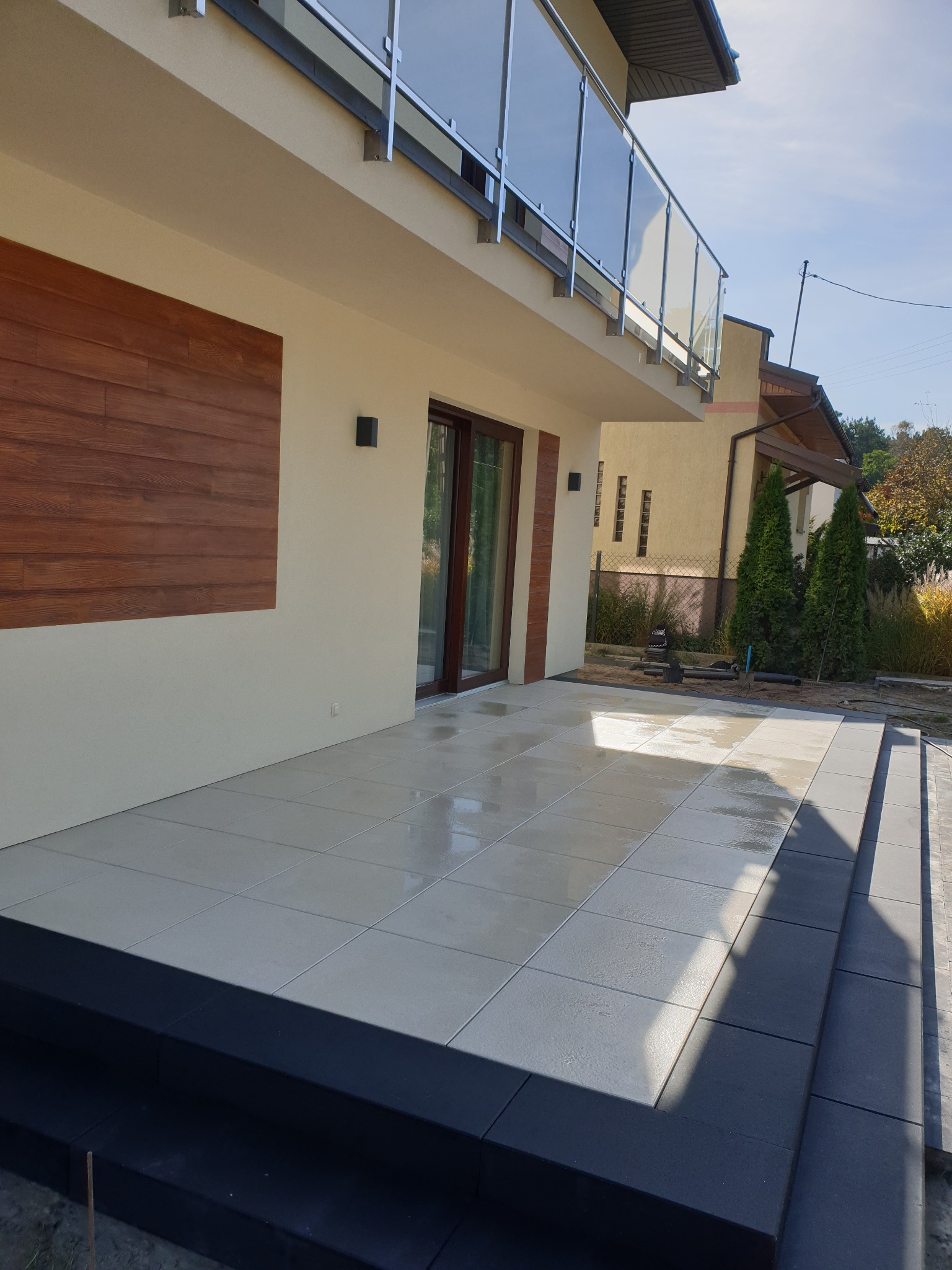 Nowoczesny taras wyłożony jasnymi płytami ceramicznymi z ciemnymi obrzeżami i schodami, przylegający do budynku z drewnianą okładziną, balkonem ze szklaną balustradą i widokiem na ogród.