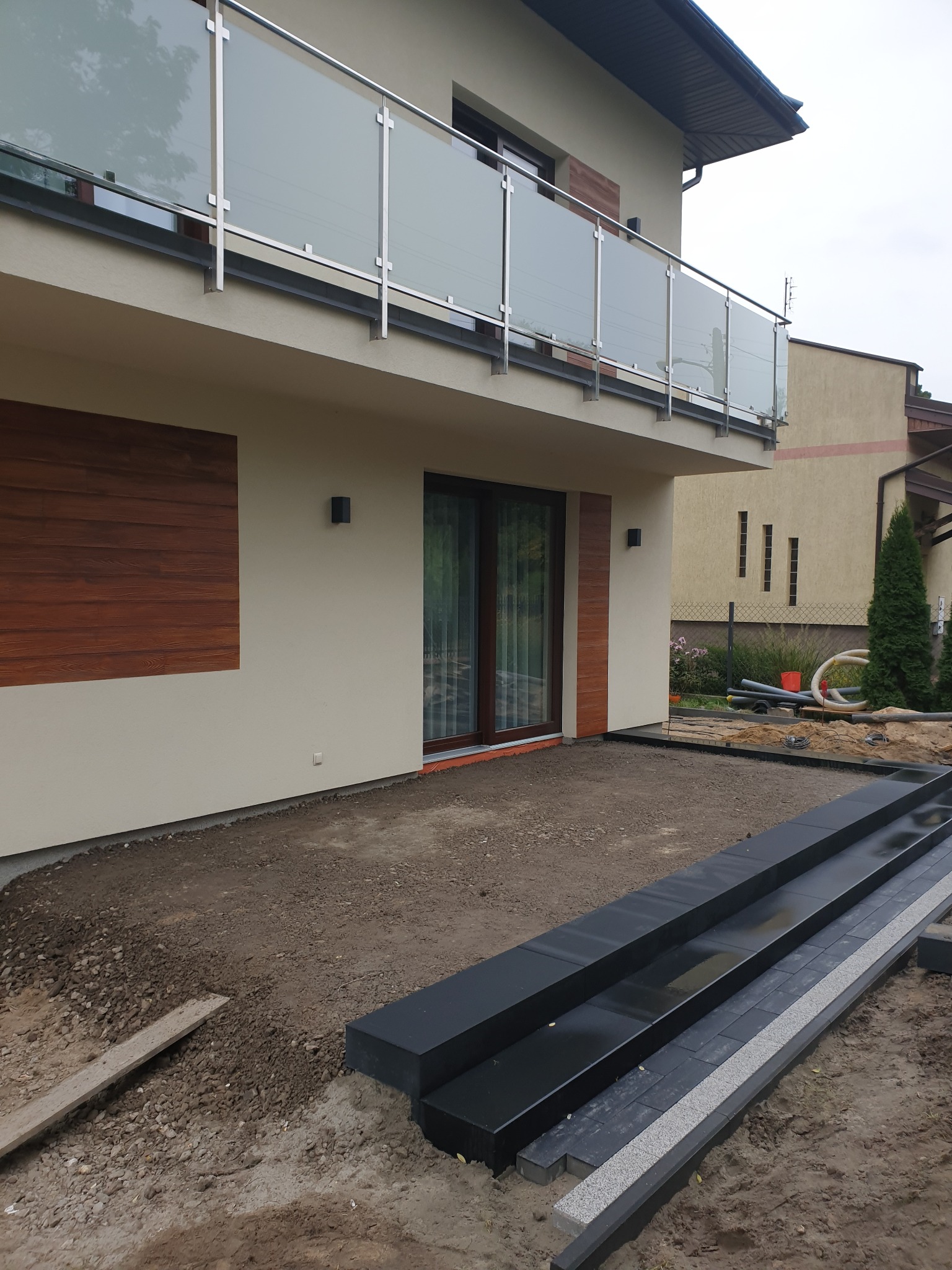 Przygotowanie podłoża pod taras z czarnych płyt kamiennych i szarej kostki obok nowoczesnego domu z balkonem ze szklaną balustradą.